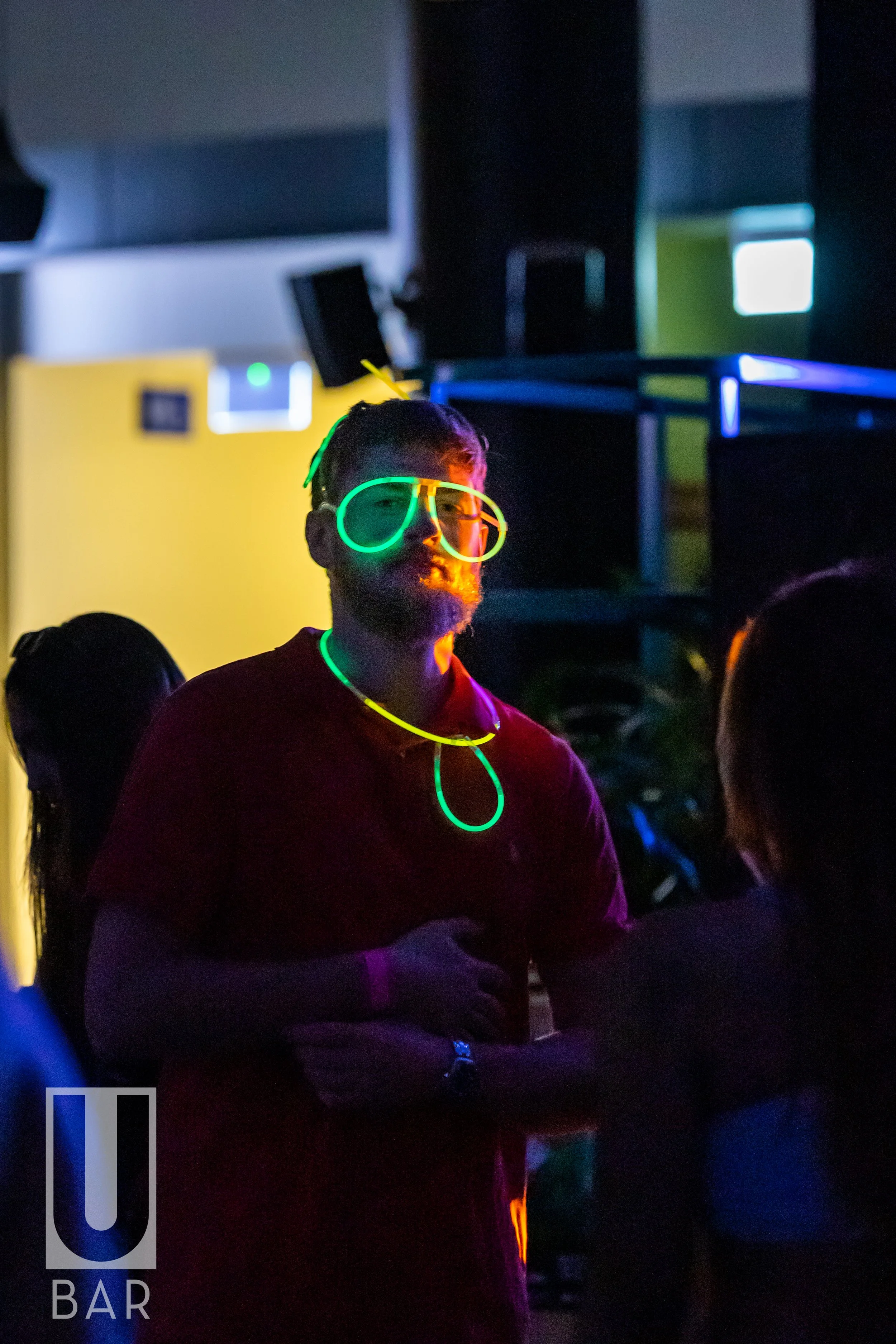 A man wearing glowing neon glasses, a glow-in-the-dark necklace, and a glow-in-the-dark bracelet stands in a dimly lit bar with other people around him. The bar has neon lighting and a modern interior.