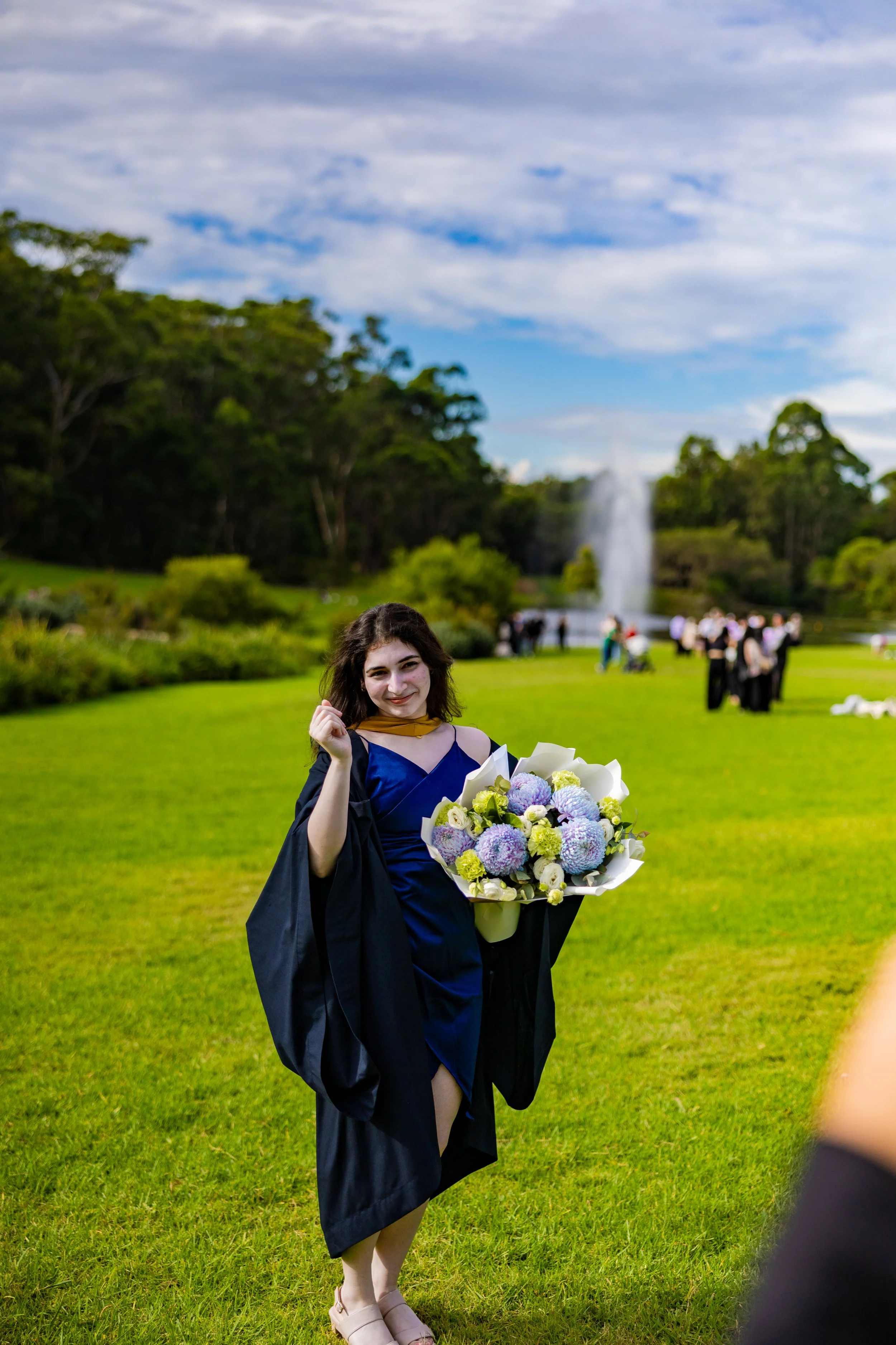 A young woman in graduation attire holding a bouquet of flowers in a park.