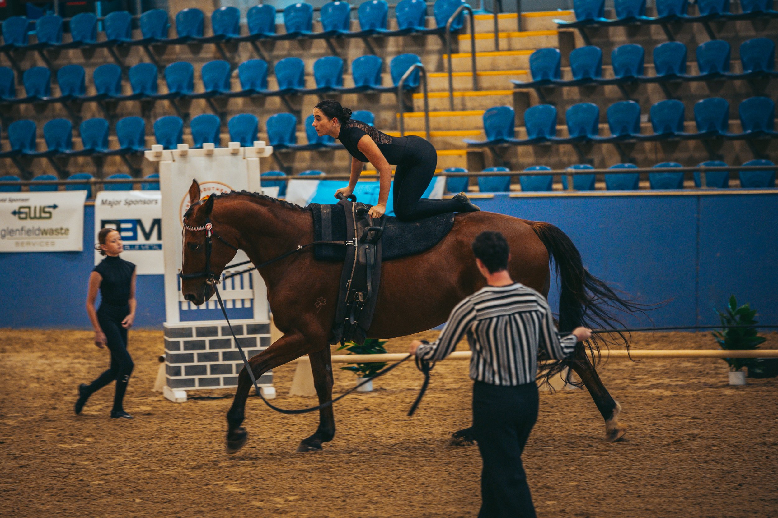 A woman is riding a horse during an indoor equestrian event, with two assistants nearby on the arena.