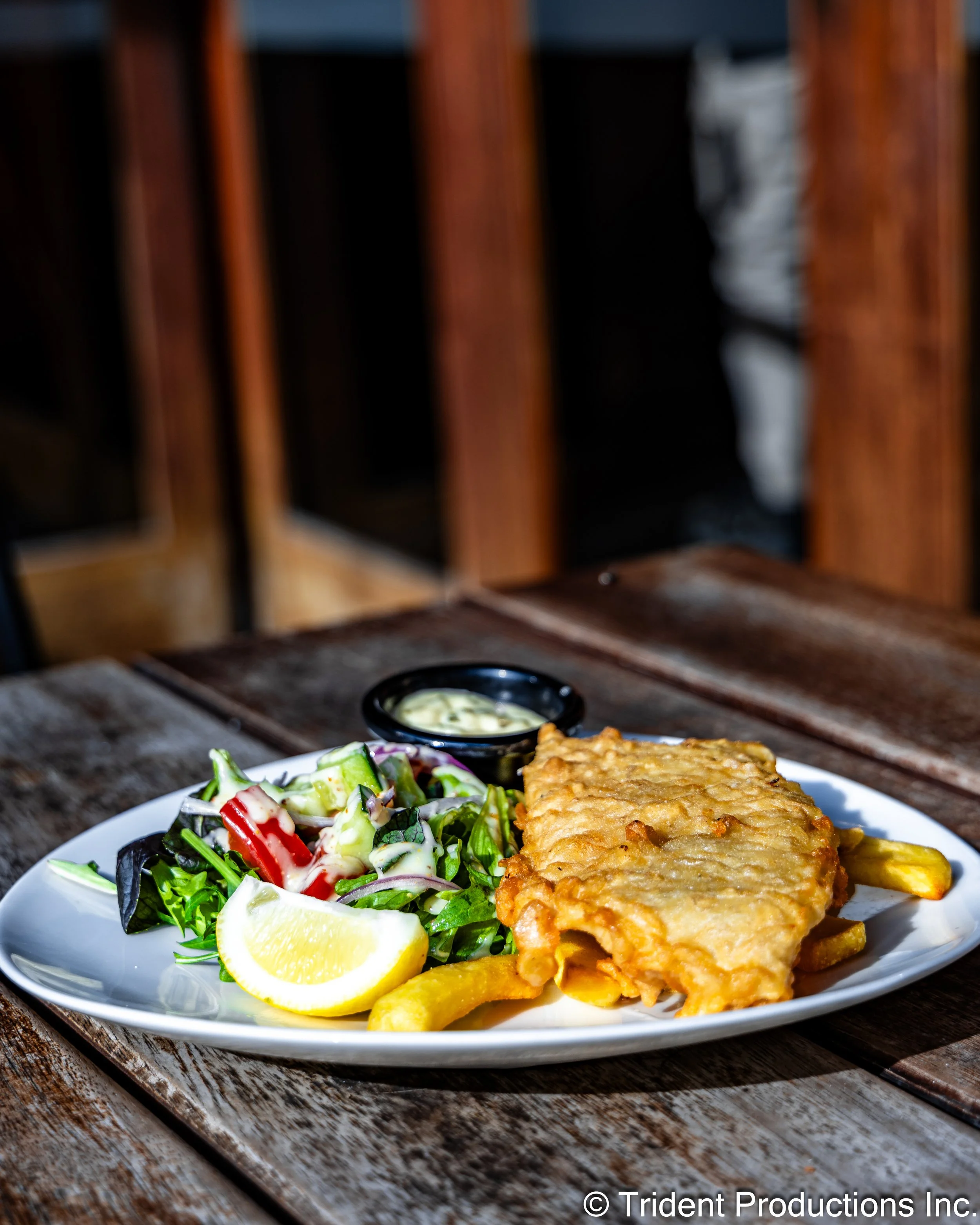 Plate with fried fish, French fries, small salad, lemon wedge, and dipping sauce on wooden table.