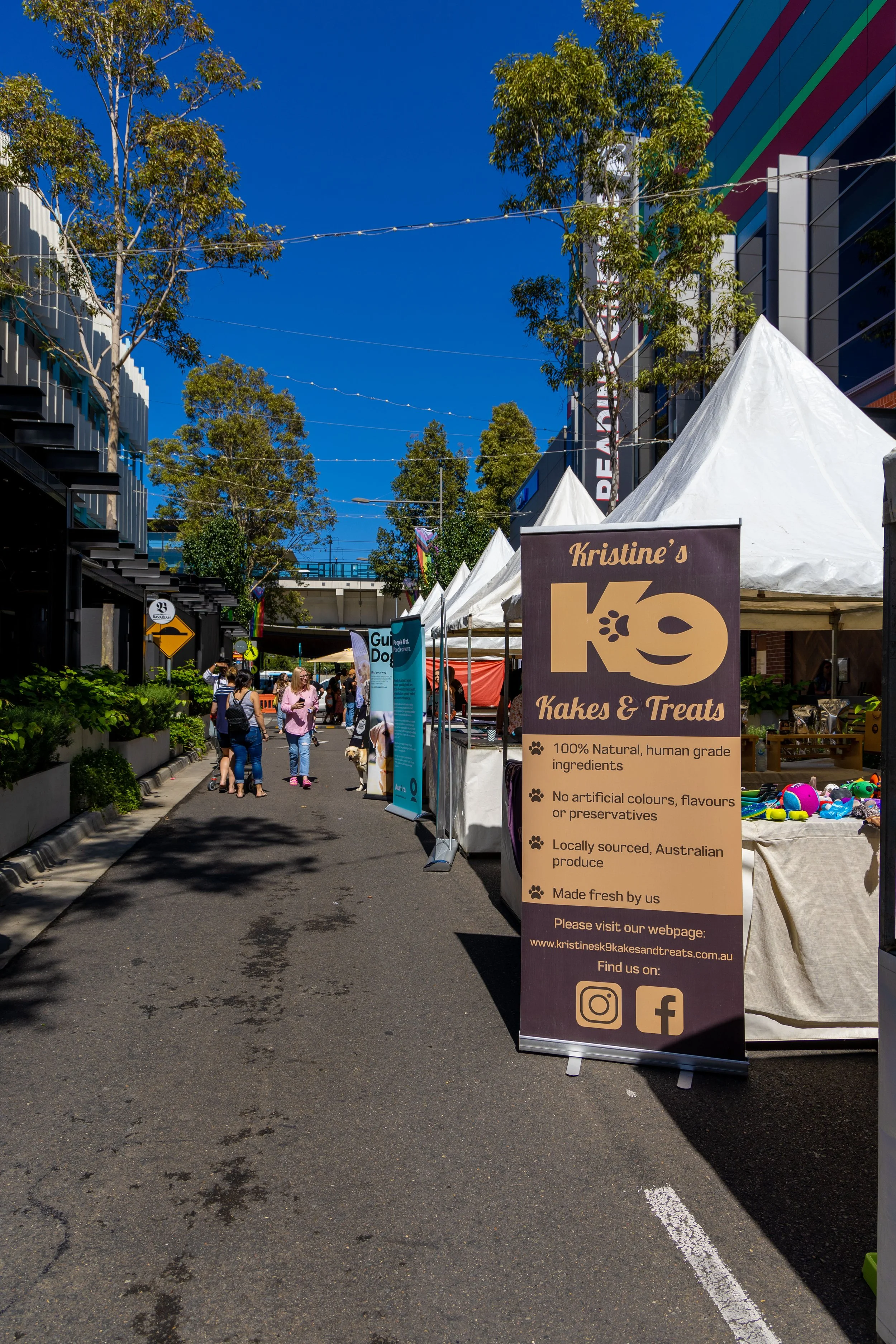Outdoor market with vendor tents and a sign for Kristine's K9 Kakes & Treats, on a sunny day with a clear blue sky, trees, and people walking and browsing.