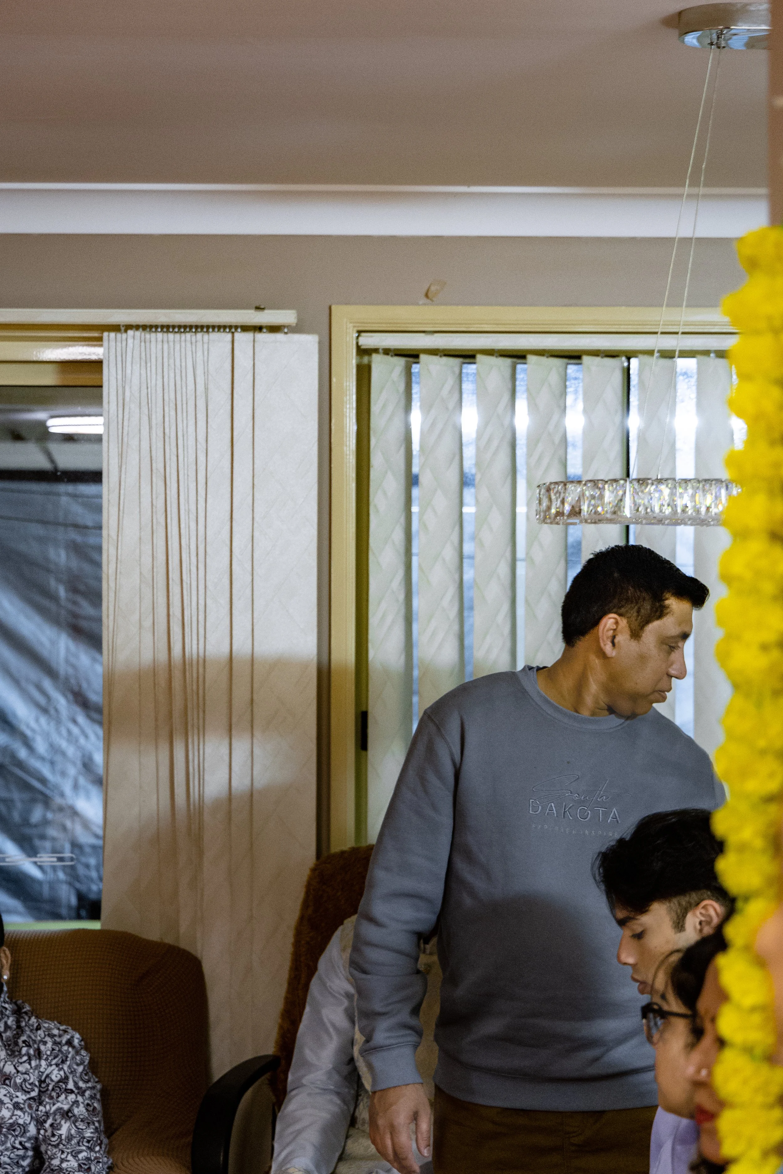 A man with dark hair wearing a gray sweatshirt standing in an indoor space with part of a woman wearing glasses and a white shirt visible in the lower right corner, yellow flowers on the right side, and a chandelier overhead.