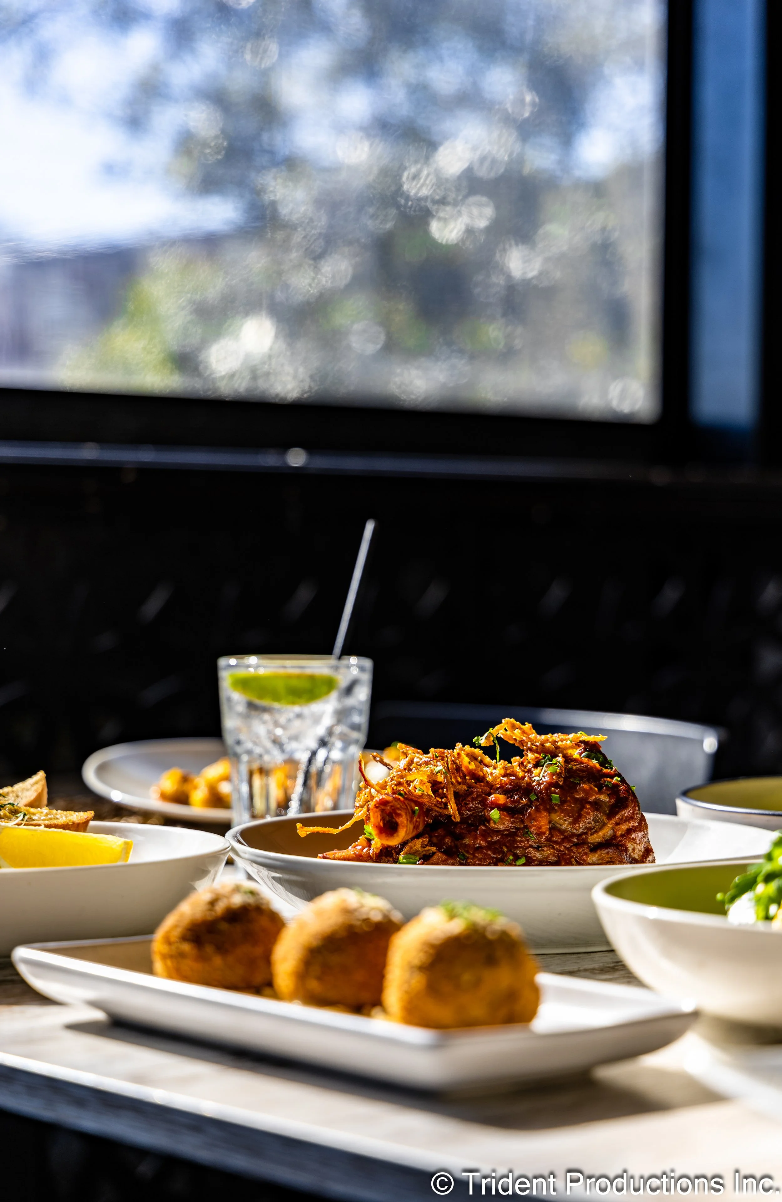 A close-up of a table with various dishes, including a glass of water with lime, fried food, and a large main course dish, set near a window with sunlight streaming through.