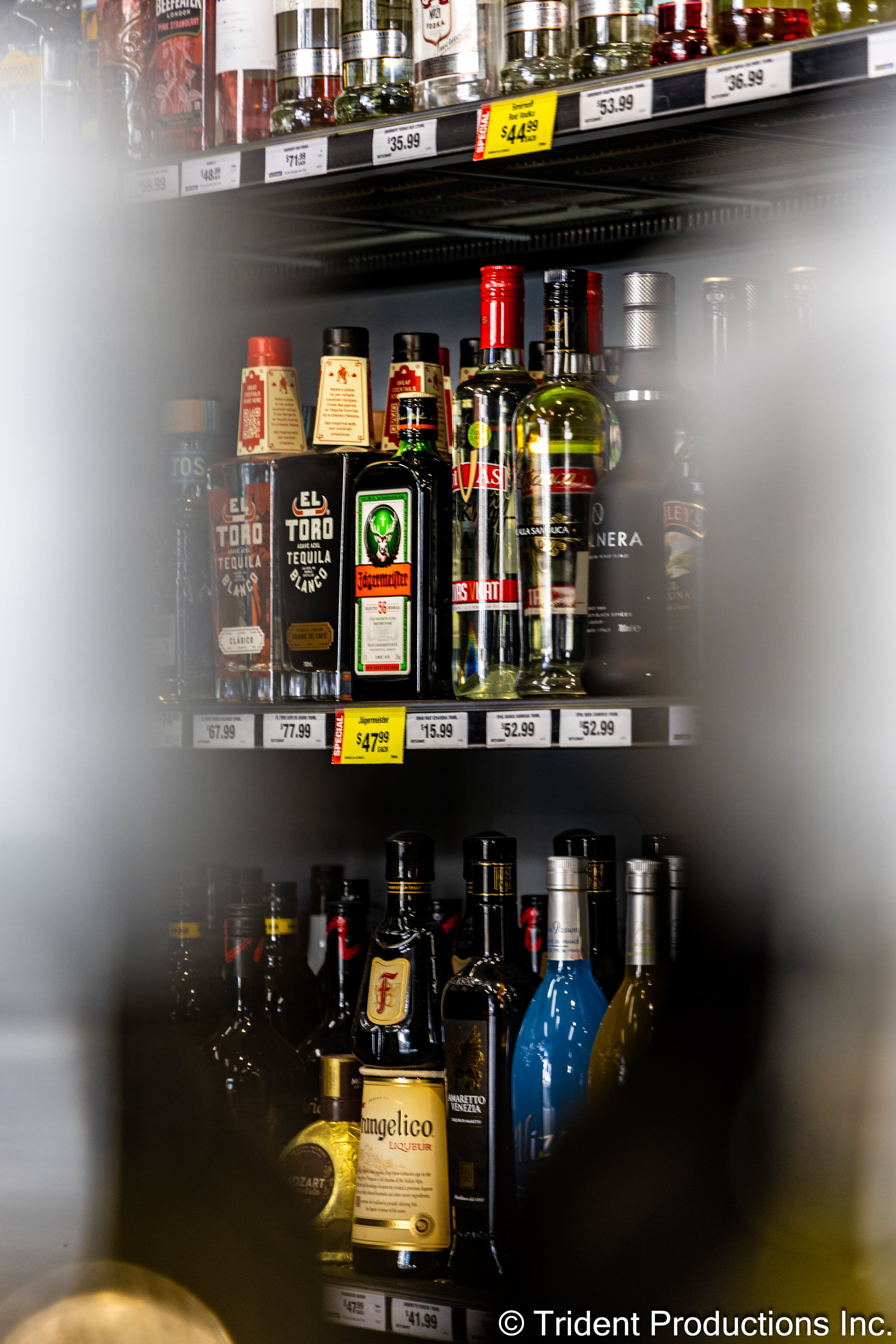 Shelves of liquor bottles in a store, including tequila, Jägermeister, and other spirits. Visible price tags and sale labels.
