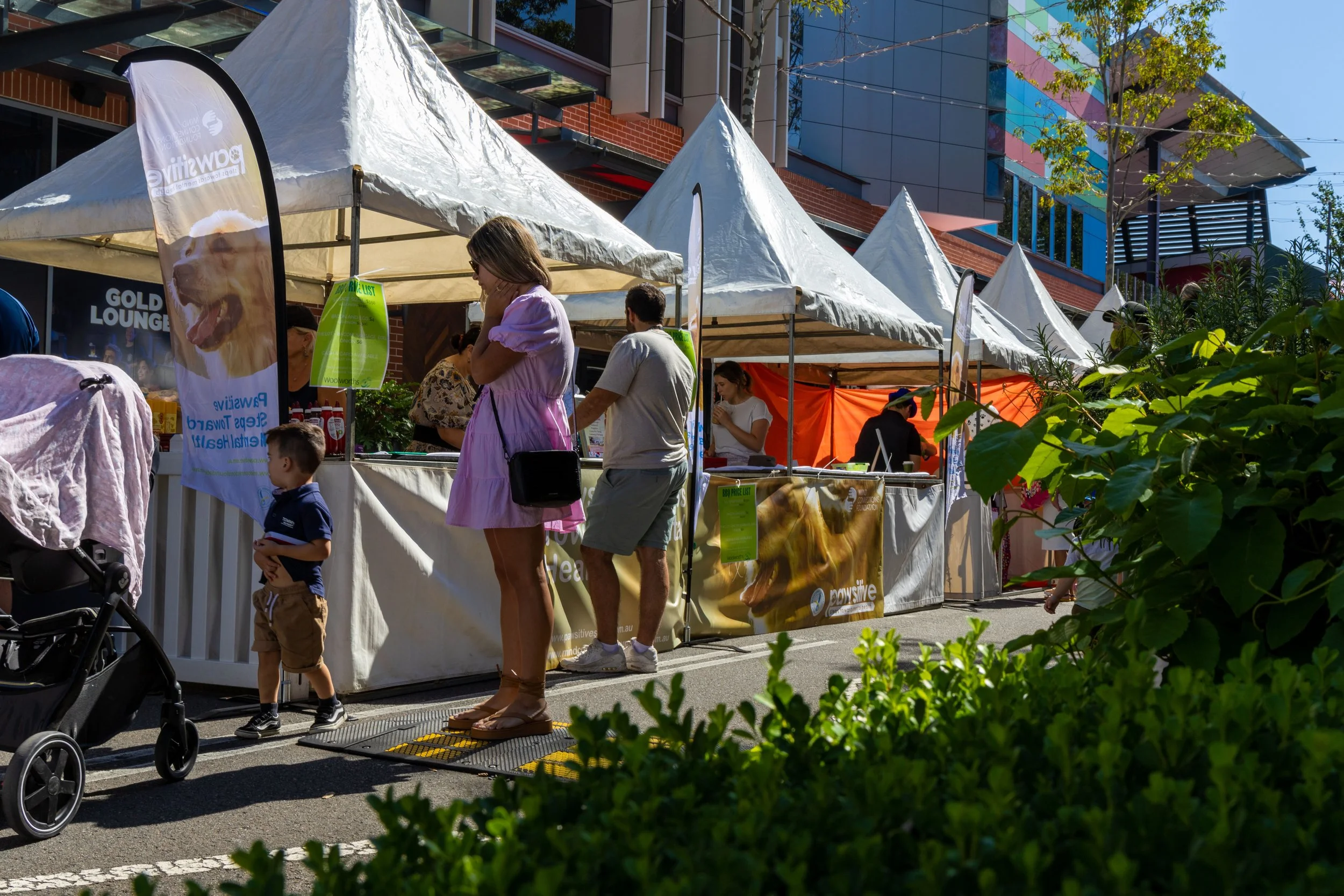 People shopping at an outdoor market with white tents on a sunny day. A woman in a pink dress and a young boy are in foreground, with others browsing various stalls in background.
