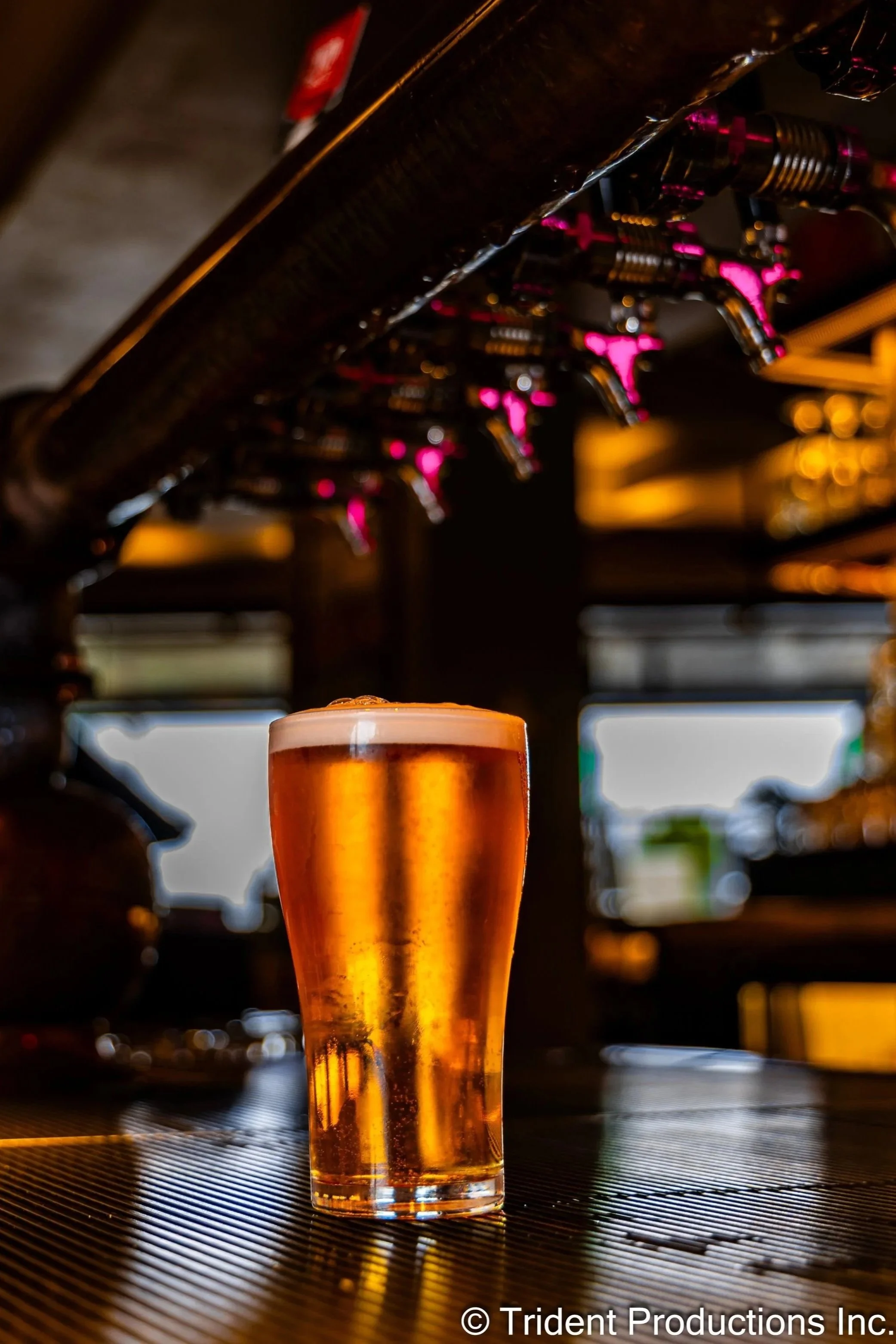 A glass of amber-colored beer with a foamy head on a bar counter, with beer taps and glowing pink lights in the background.