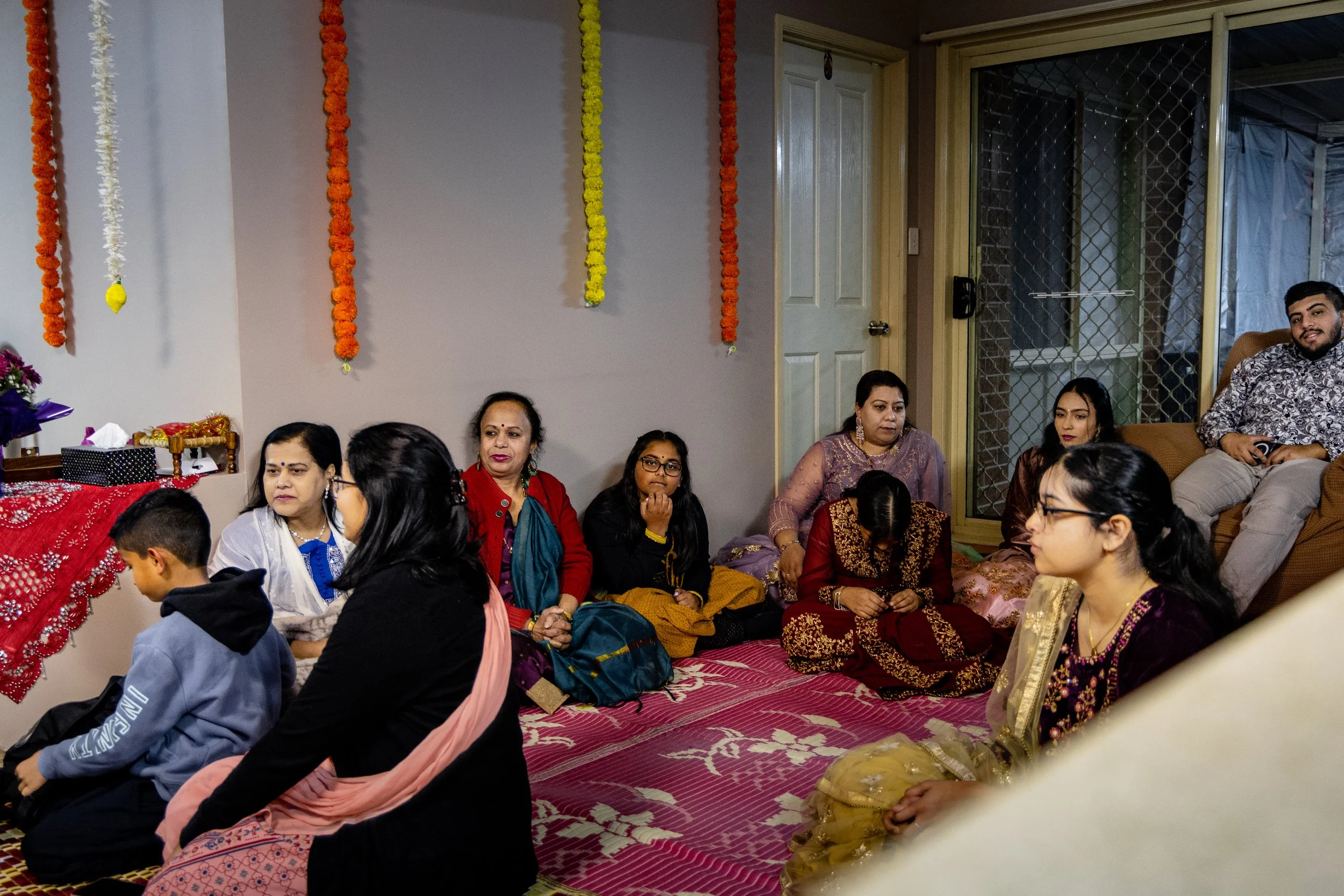 A group of people gathered in a living room for a celebration, seated on a pink patterned carpet with festive decorations hanging on the wall.