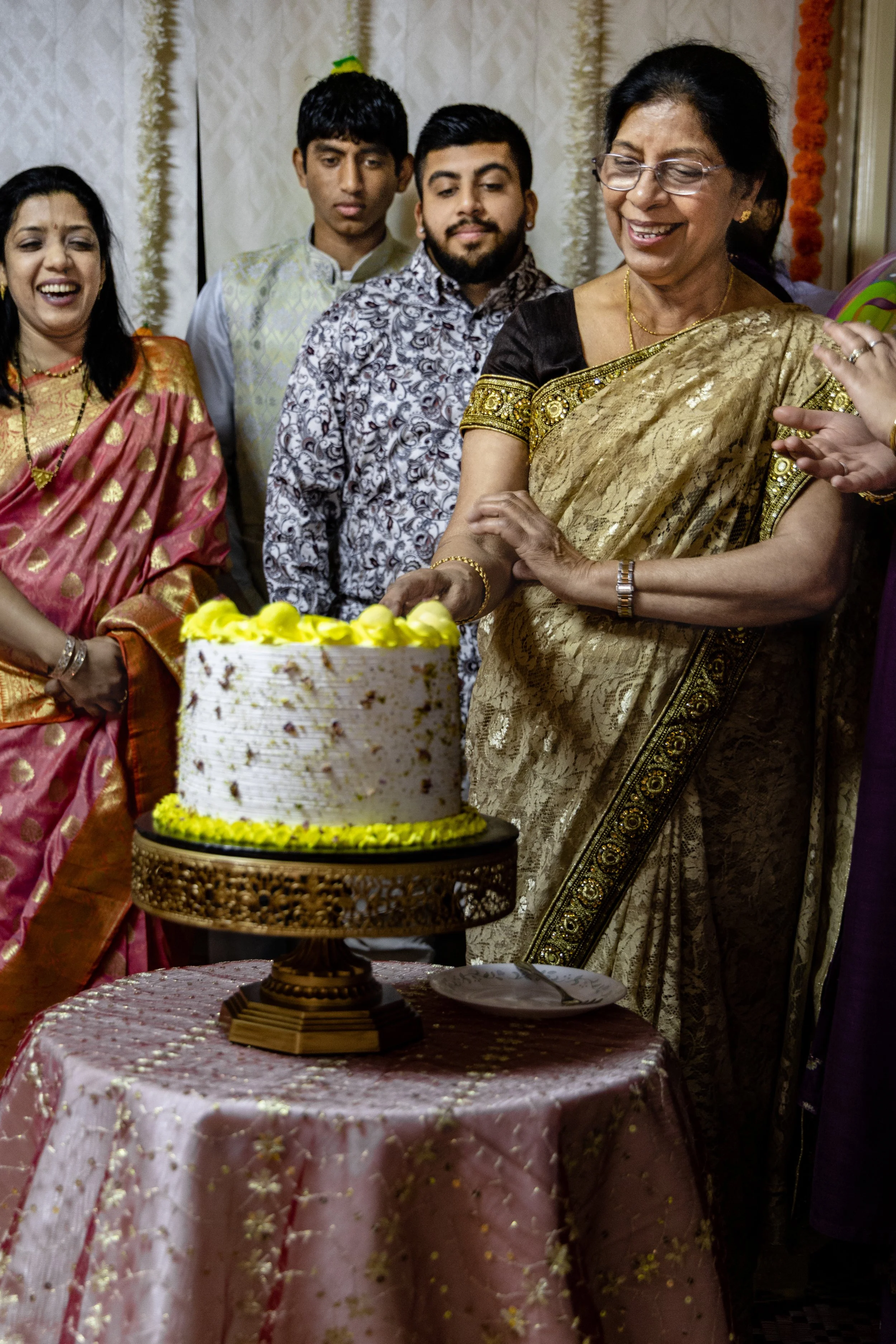 A group of people celebrating around a cake at a festive gathering. A woman in a gold and black saree is cutting the cake, smiling. Others stand close, watching and smiling. The table has a pink, embroidered cloth. The background features cream-color