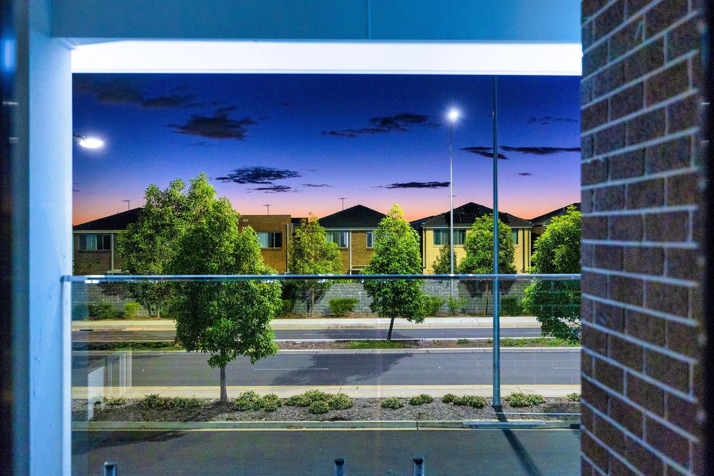 View of a suburban street at dusk, framed by a window with brick wall on the right and a blue window frame on the left, with trees, and residential houses in the distance under a colorful evening sky.