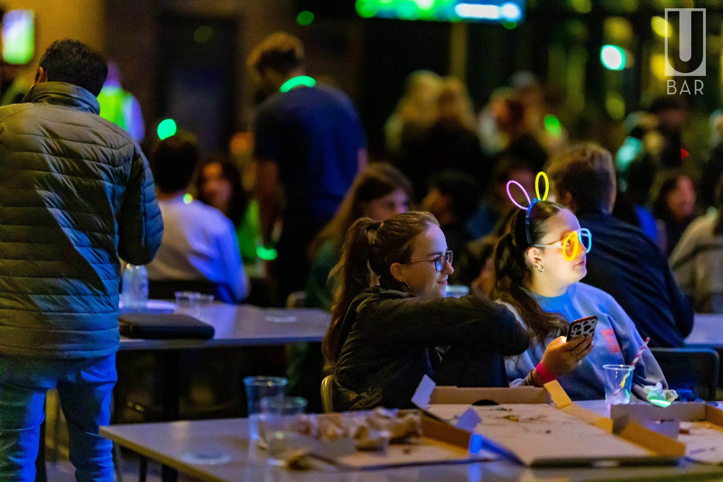 People sitting at a bar with some wearing glowing glasses and bunny ears, enjoying drinks and socializing at night.