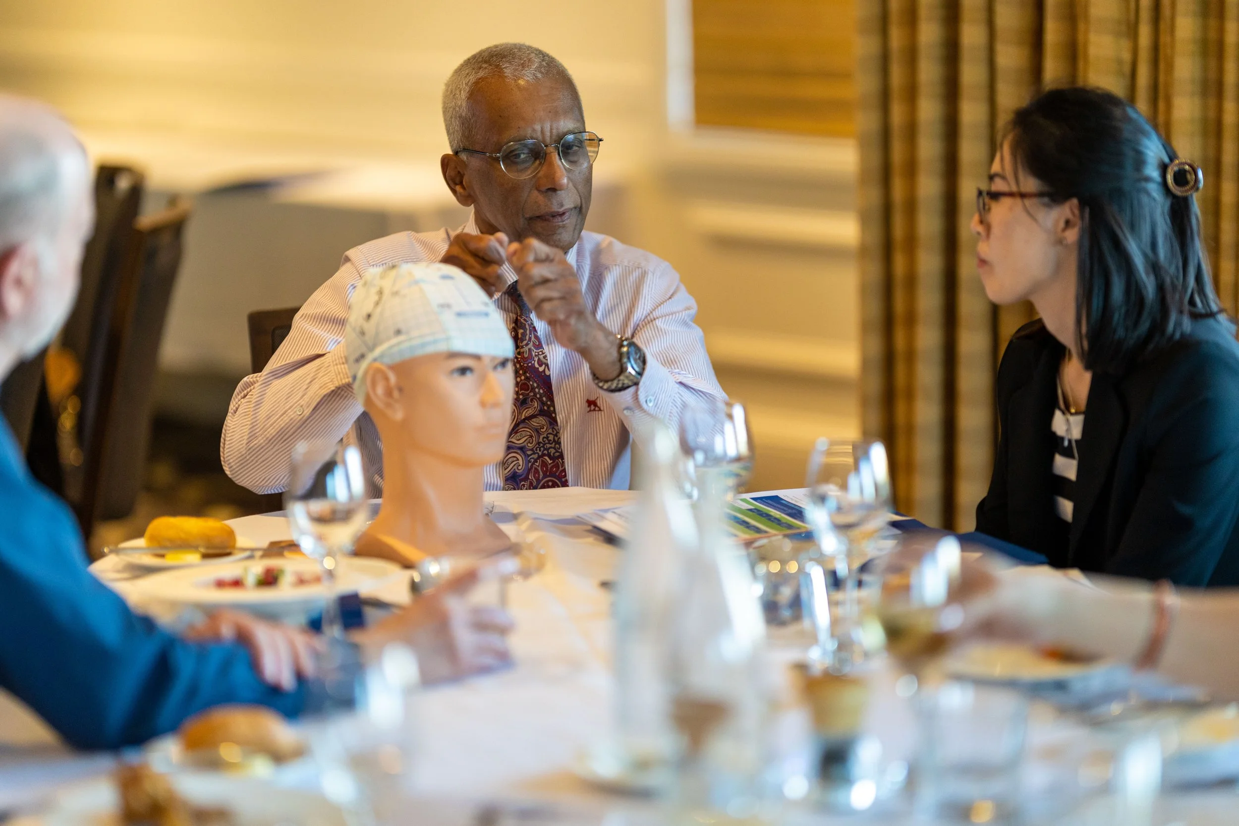A group of people seated at a dining table during a discussion. One man is talking, with a mannequin head on the table in front of him, and others are listening. The table is set with glasses and plates.