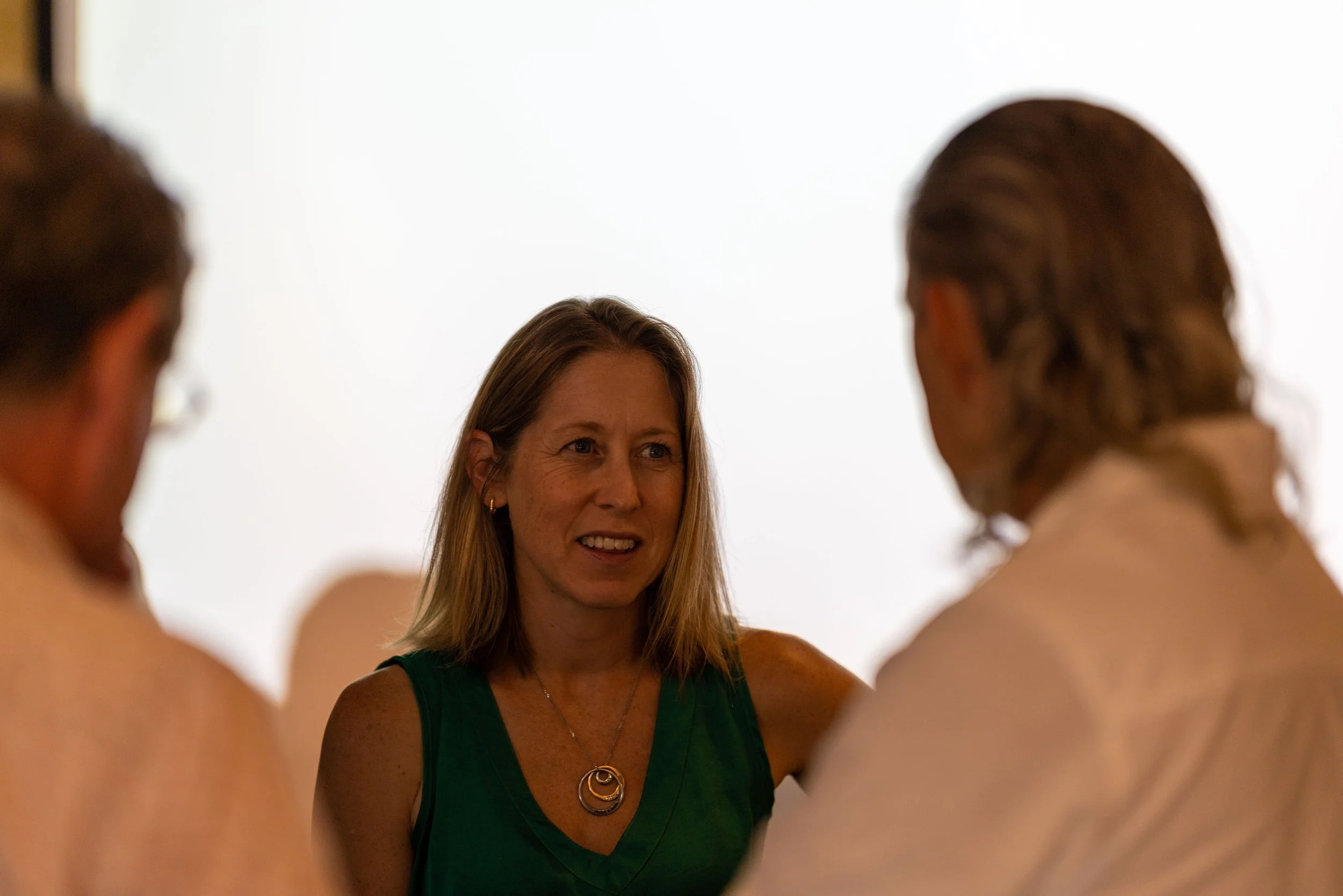 A woman in a green top speaking to two men with long hair, in a social or professional setting with a plain white background.