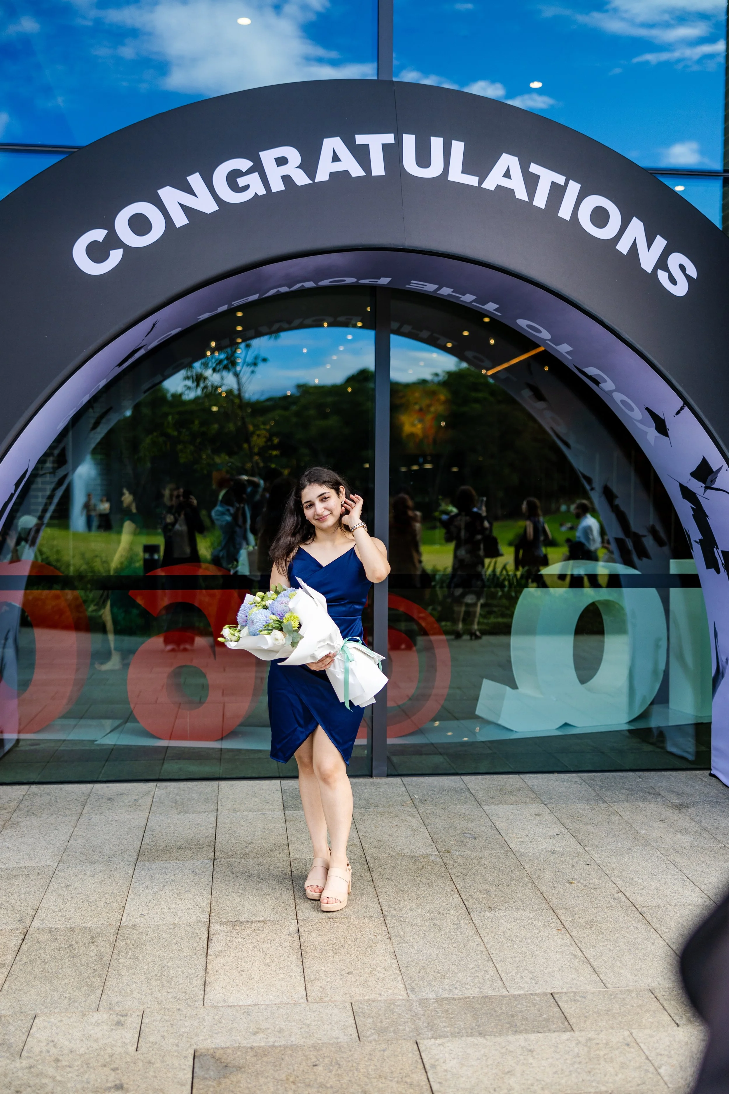 Young woman in a blue dress holding a bouquet of flowers, standing in front of a building with a sign that reads 'CONGRATULATIONS', celebrating a graduation or achievement.