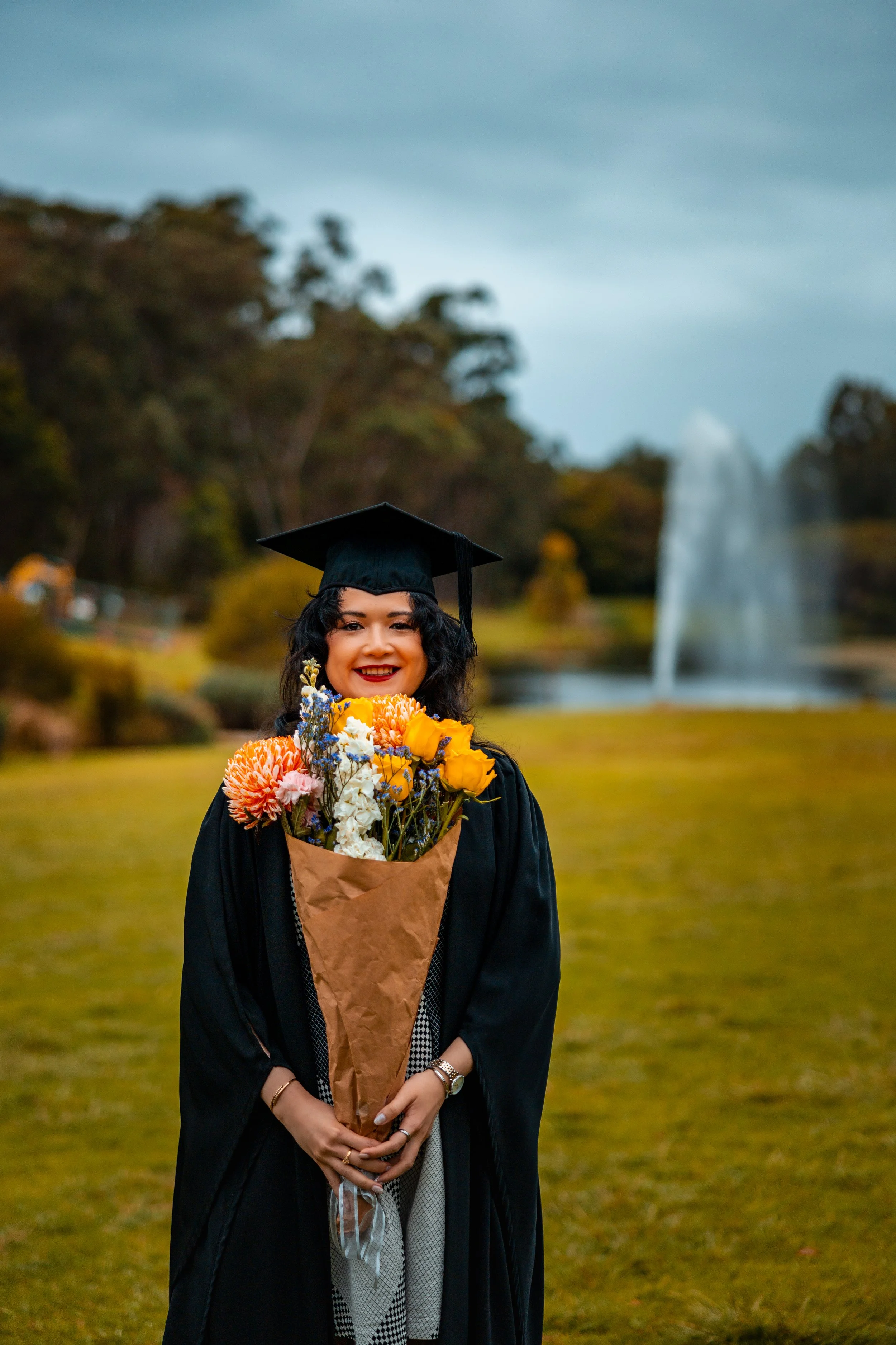A graduate in a black gown and mortarboard holding a bouquet of flowers outdoors with a fountain and trees in the background.