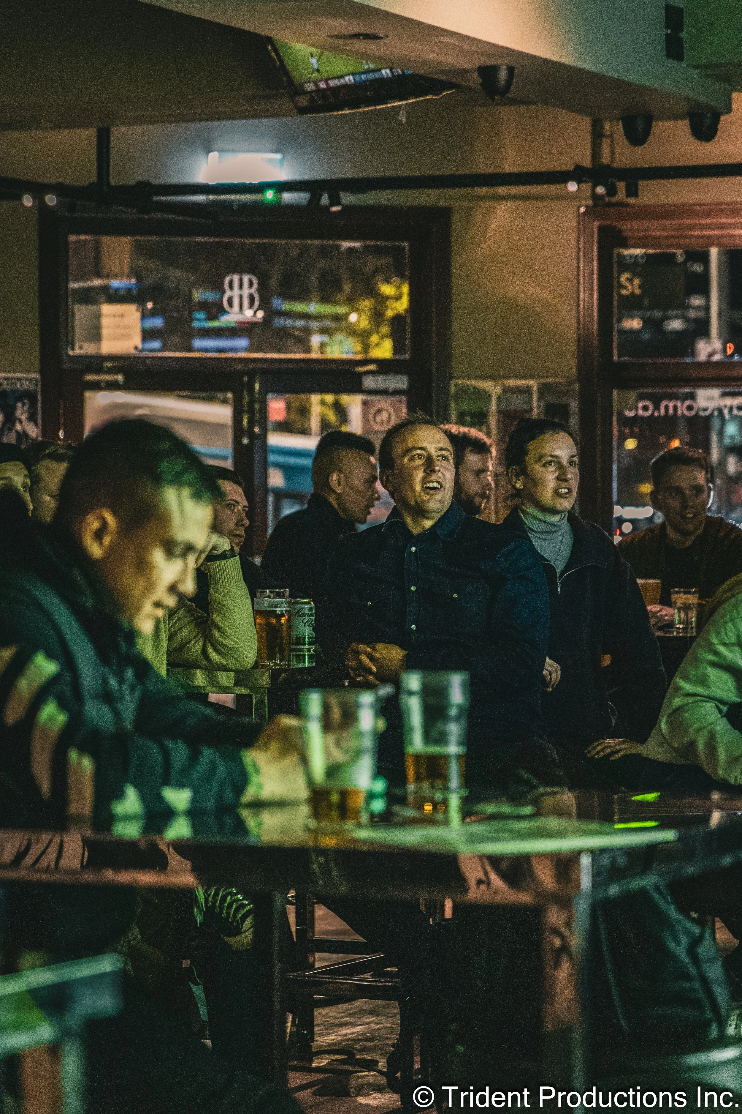 People sitting at tables in a bar or pub, watching a TV screen, some with drinks in front of them.