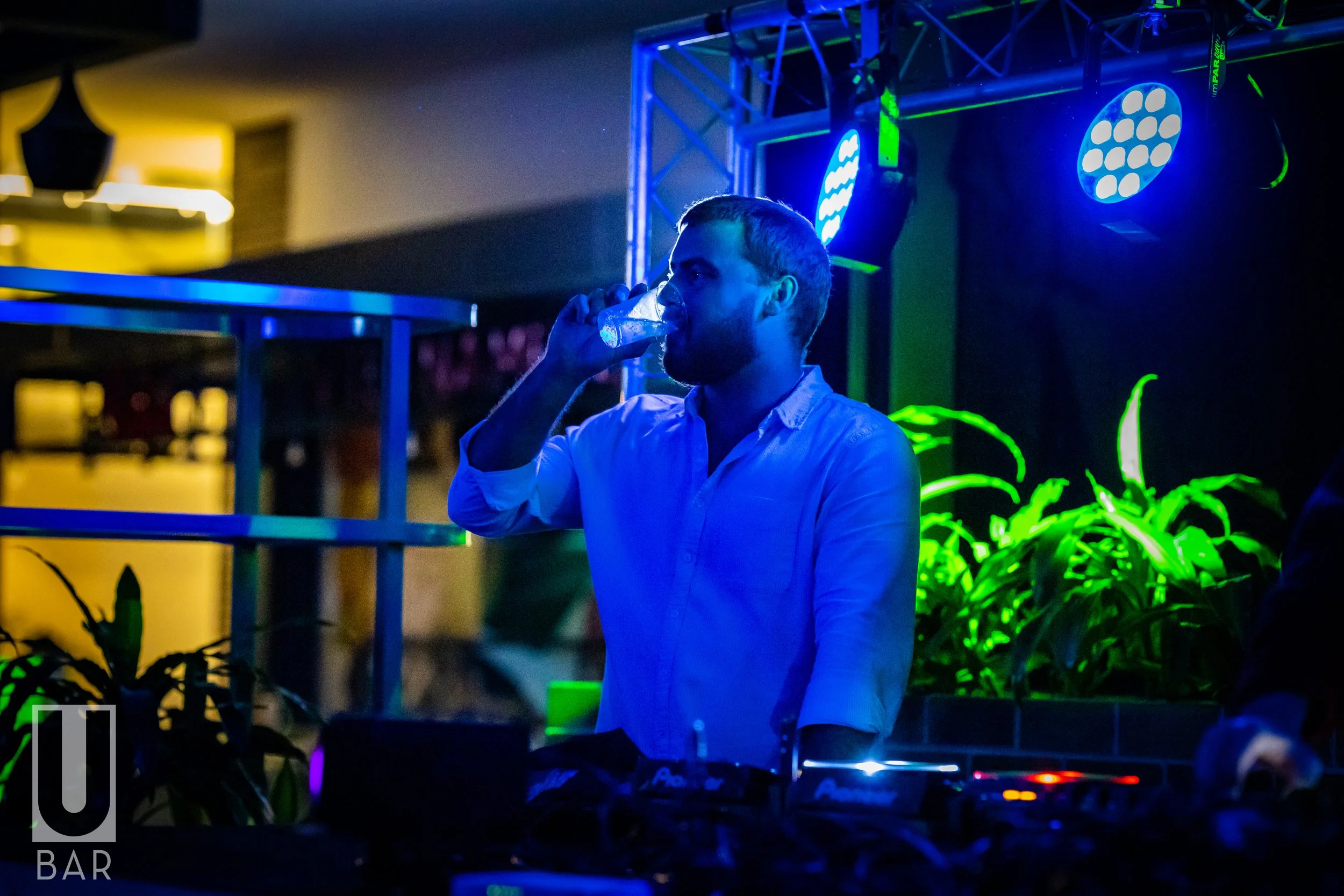 A man in a white shirt drinking from a clear glass at a nightclub or bar, illuminated by blue and green LED lights with DJ equipment visible in the foreground.