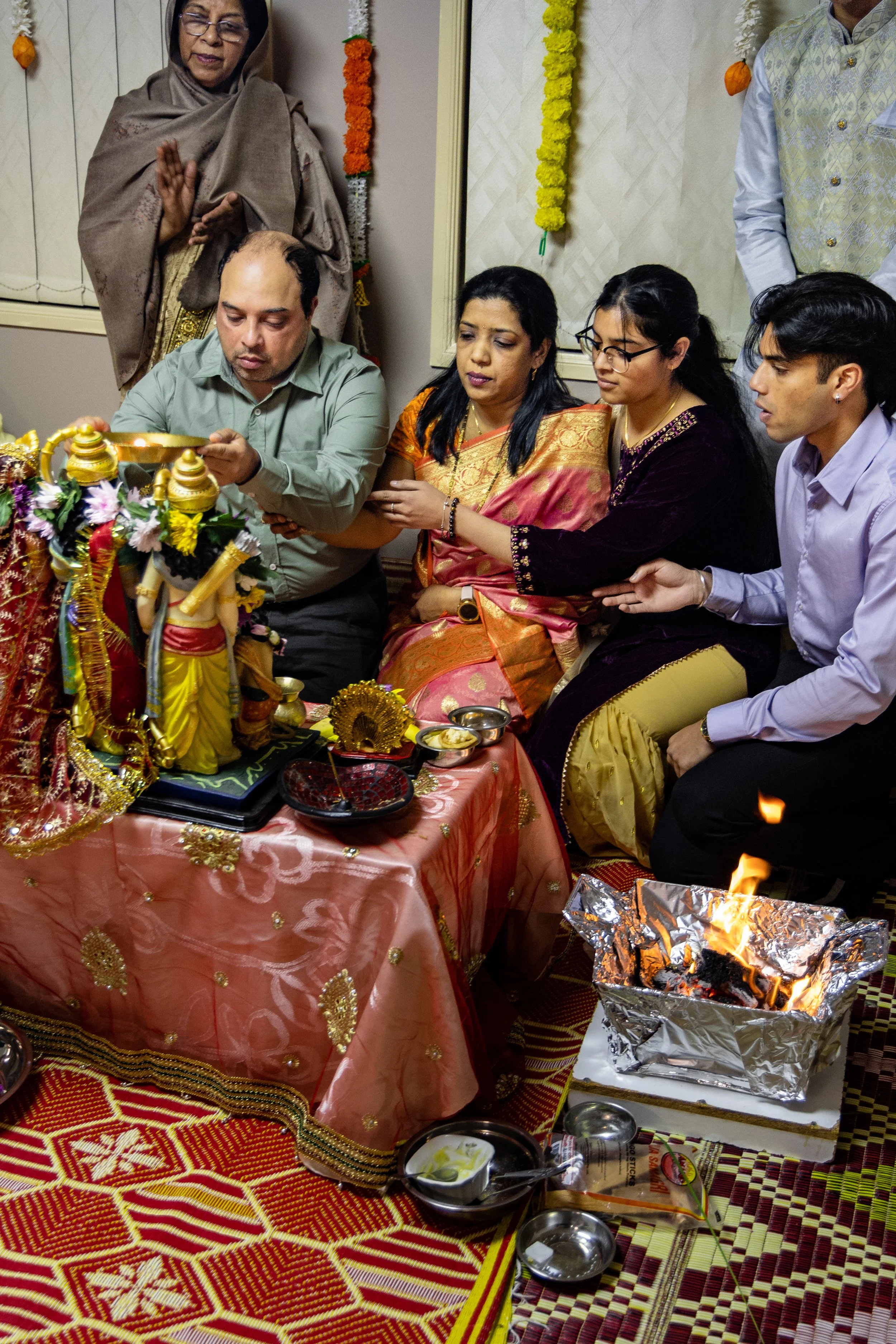 People participating in a traditional Indian religious ceremony, sitting around a decorated altar with statues, flowers, and offerings, as a fire burns in a metal container.