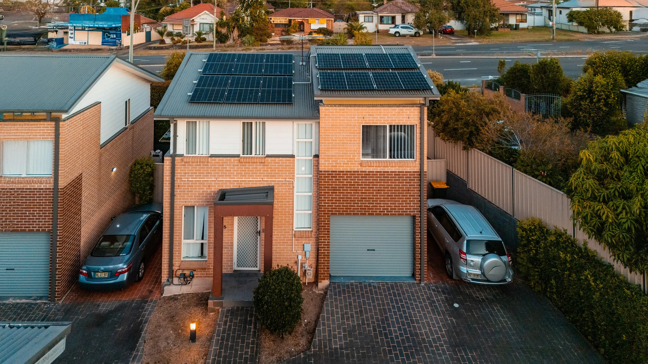 An aerial view of a modern two-story brick house with solar panels on the roof, driveway, surrounding greenery, and neighboring houses in the background.