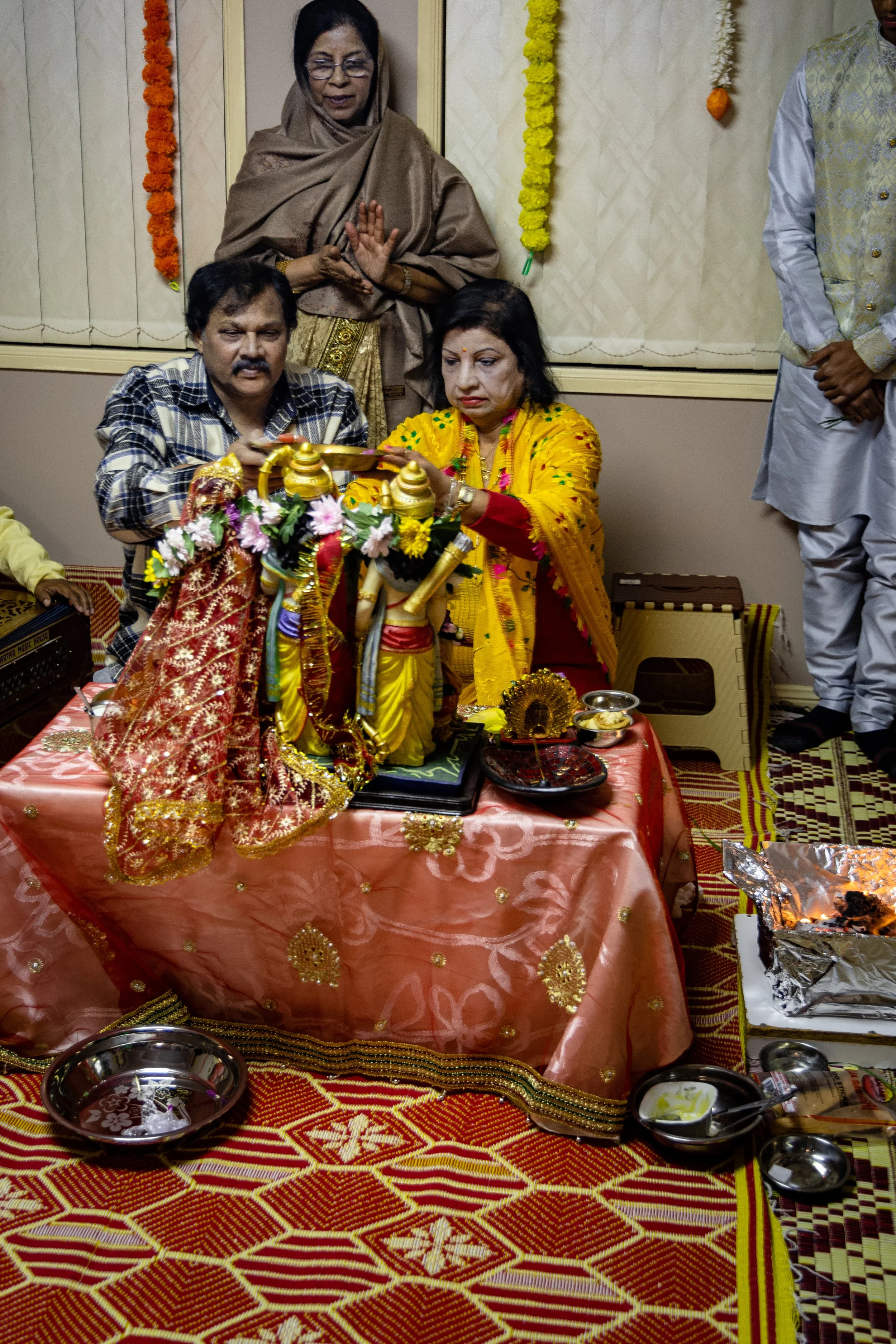 A group of people participating in a Hindu religious ceremony. A woman in a yellow saree is performing a ritual in front of a decorated idol of a deity, with others observing. The scene includes ritual items and offerings on a decorated table and flo