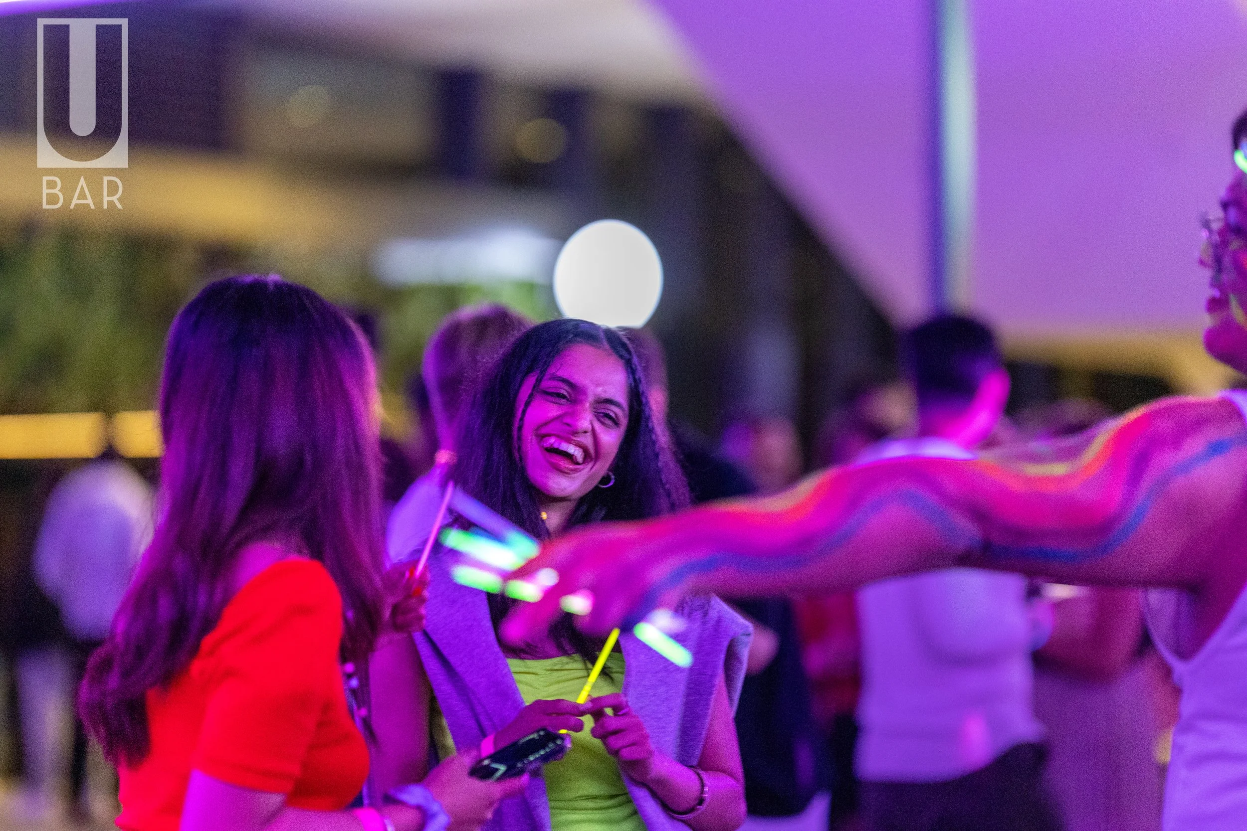 People enjoying nightlife at a bar, illuminated with purple and green lights, with a smiling woman holding glow sticks.
