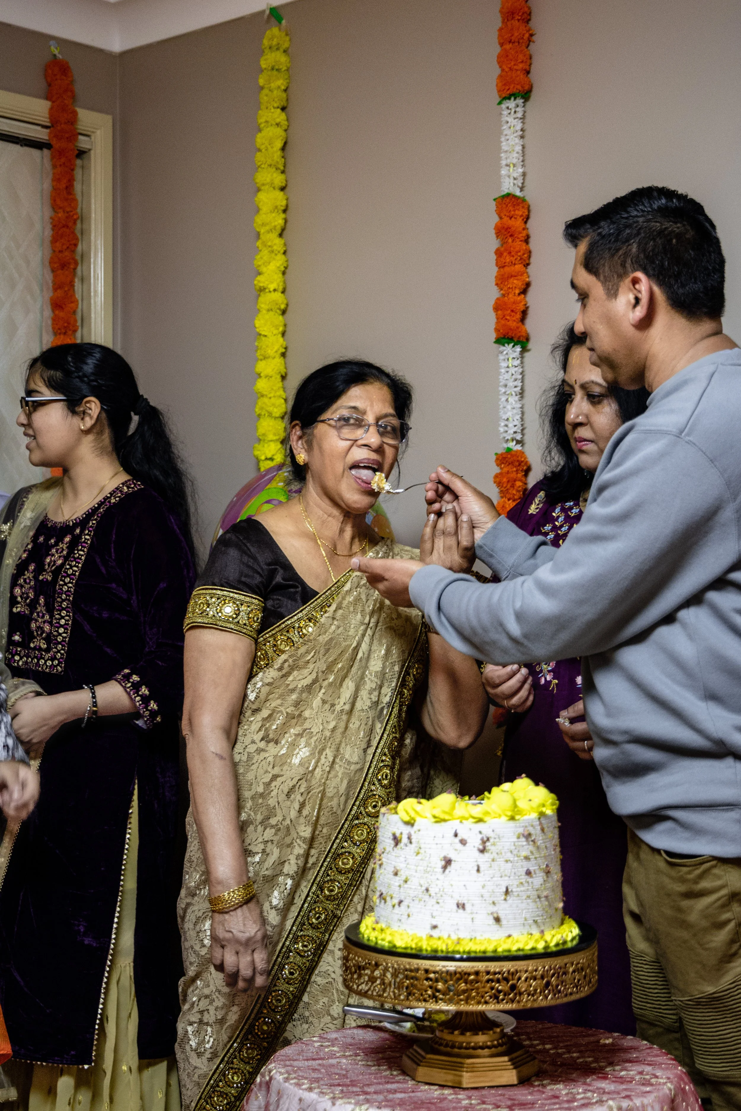 People celebrating a birthday with a cake and traditional Indian attire, surrounded by colorful floral decorations.