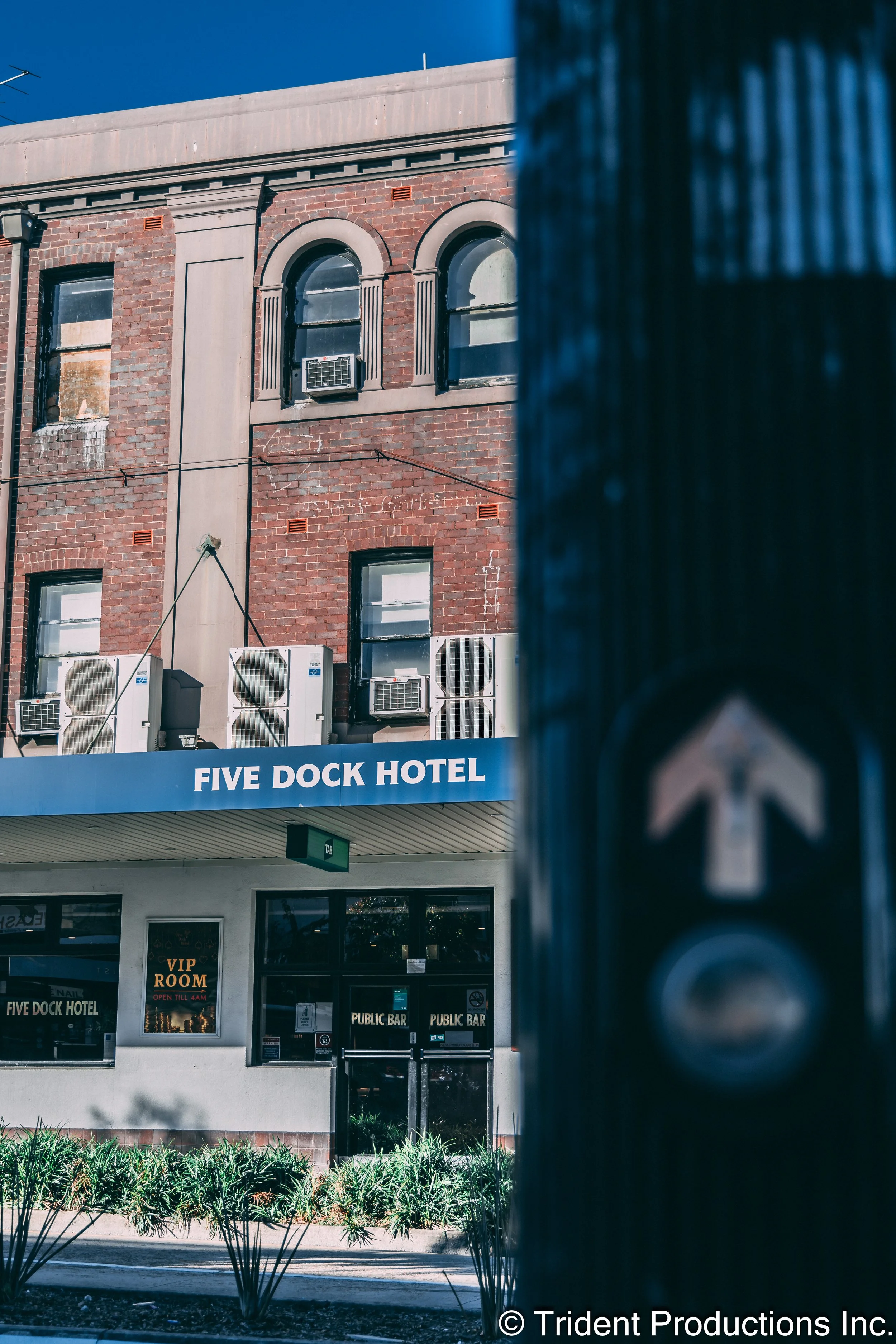 A brick building with multiple air conditioning units and arched windows, labeled "FIVE DOCK HOTEL." The ground floor has a sign for a VIP room and public bar, with glass doors and greenery outside.