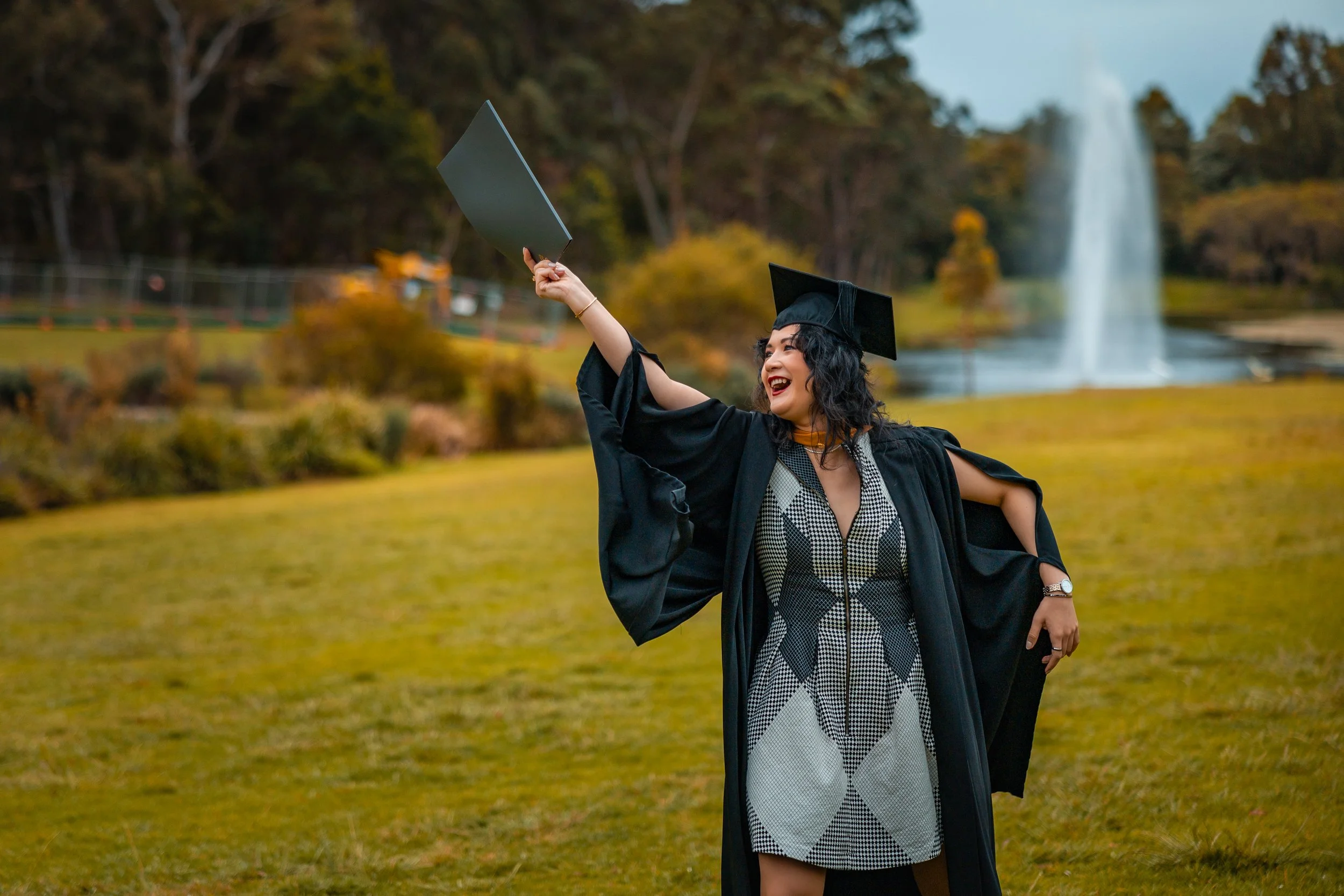 Woman in graduation cap and gown celebrating outdoors with a fountain and trees in the background