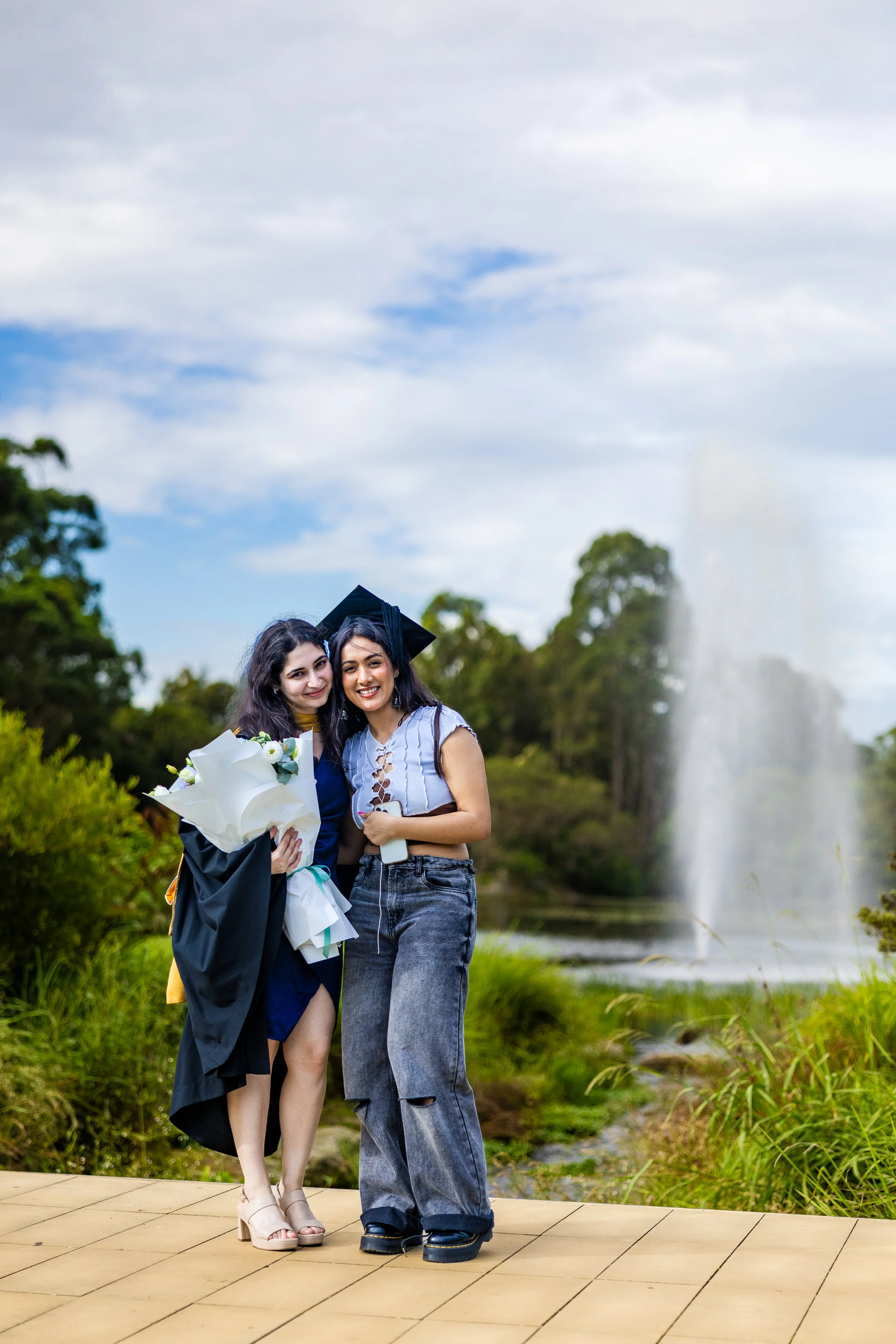 Two young women standing outdoors on a wooden platform, one wearing a graduation gown and cap, holding a bouquet of flowers, and the other in casual clothing, with a fountain and trees in the background on a partly cloudy day.