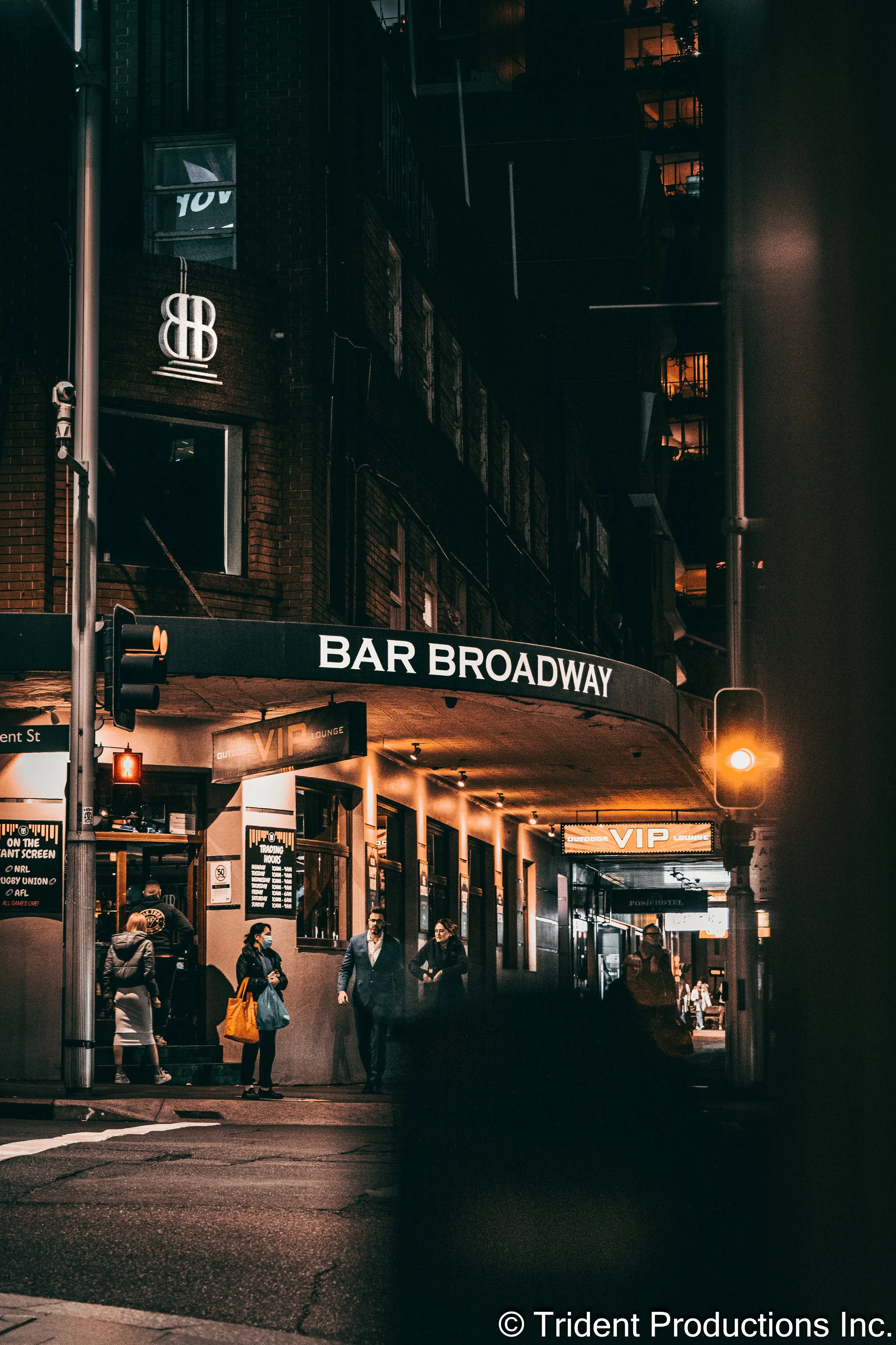 Nighttime scene outside Bar Broadway with people walking past, illuminated signs including VIP Lounge, and a building with lit windows in the background.