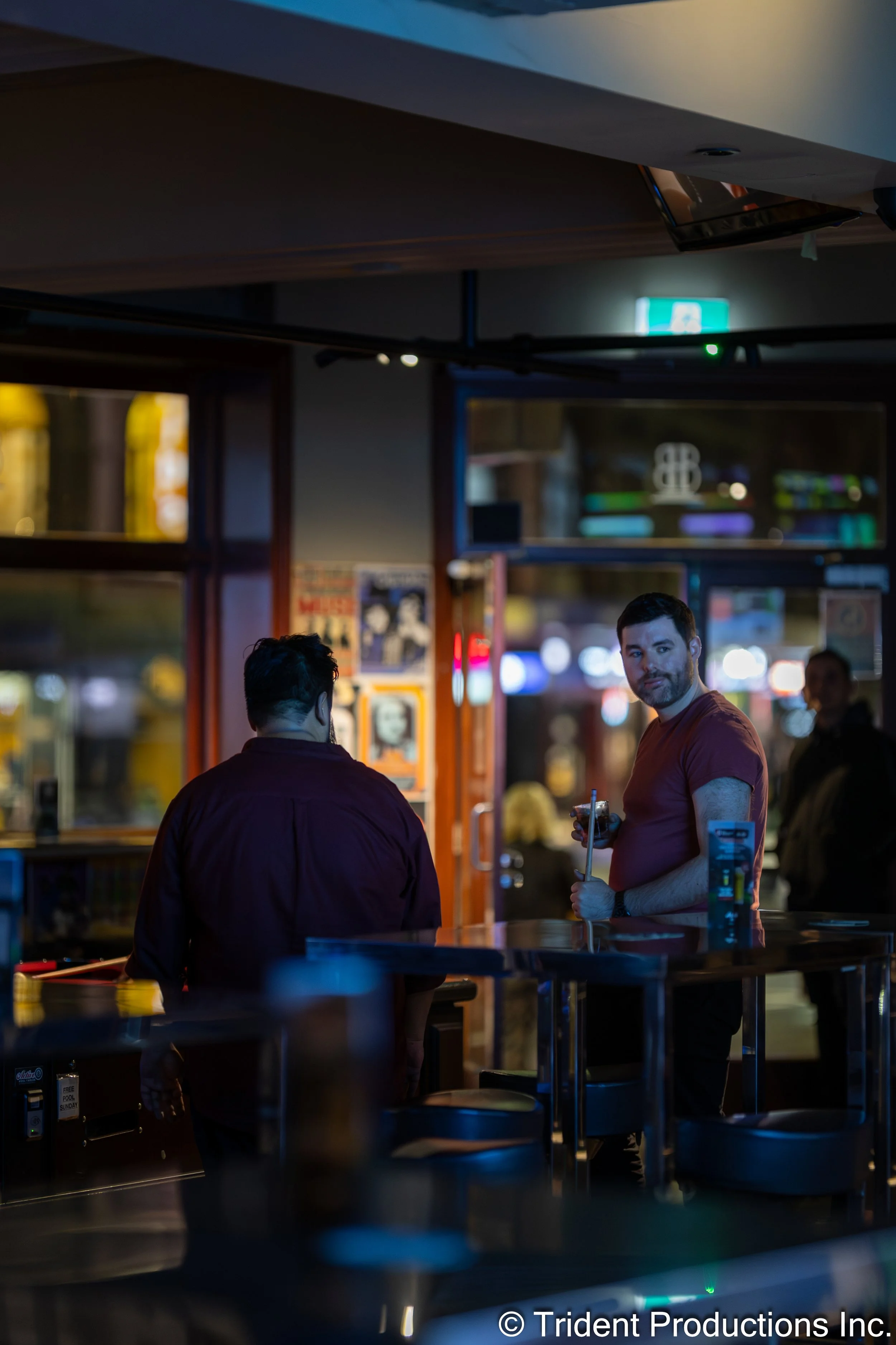 Two men inside a bar or restaurant at night, one facing away and the other looking at the camera holding a drink with a straw, with dim colorful lighting and blurred background of street lights.