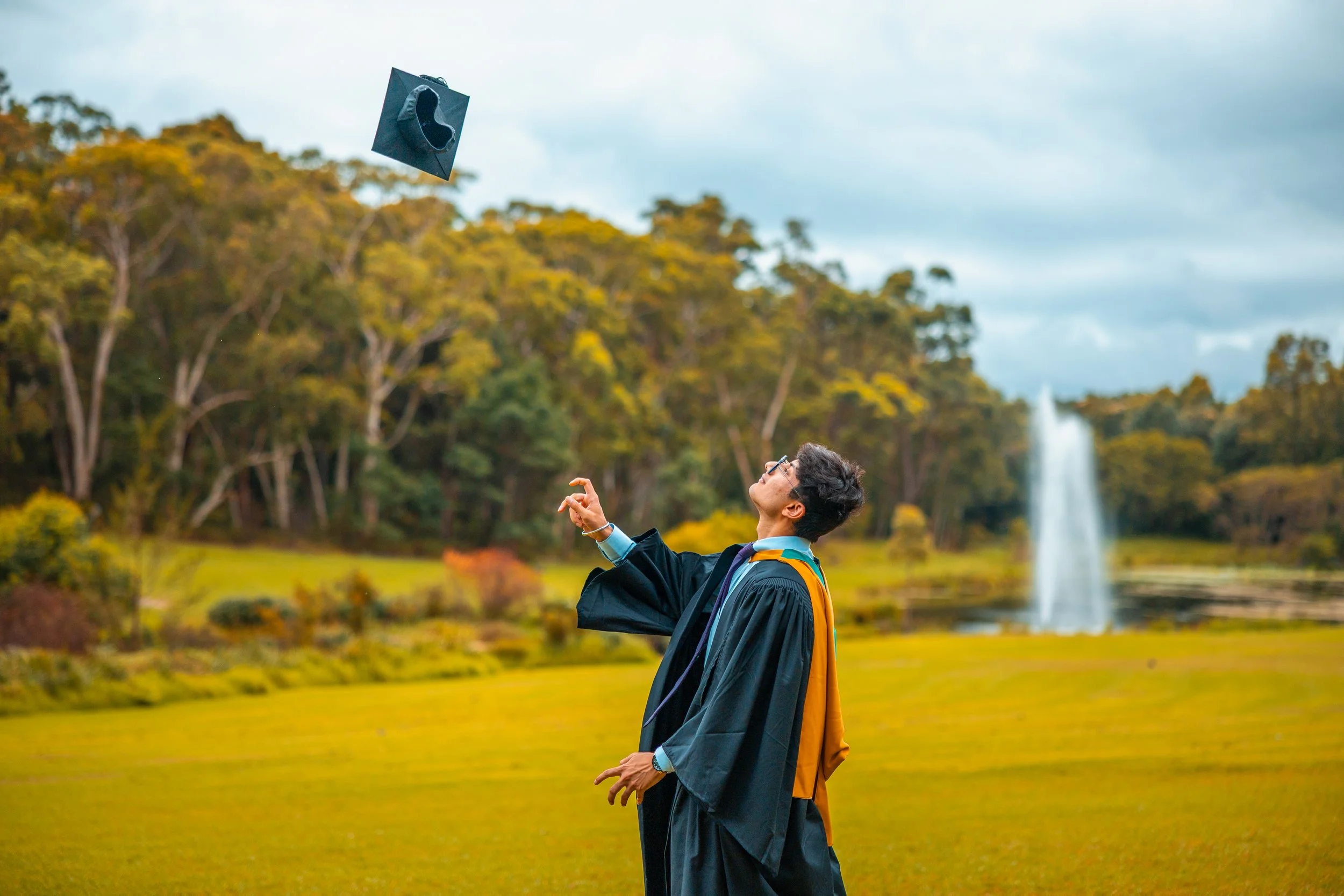 A man in graduation gown and cap tossing his cap into the air outdoors with a fountain and trees in the background.