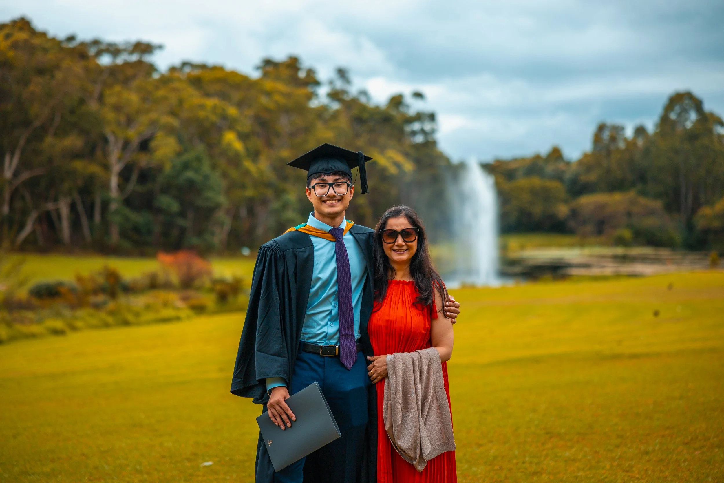 A young man in graduation gown and cap with glasses, standing next to a woman in a red dress and sunglasses, outdoors near a fountain with trees in the background.