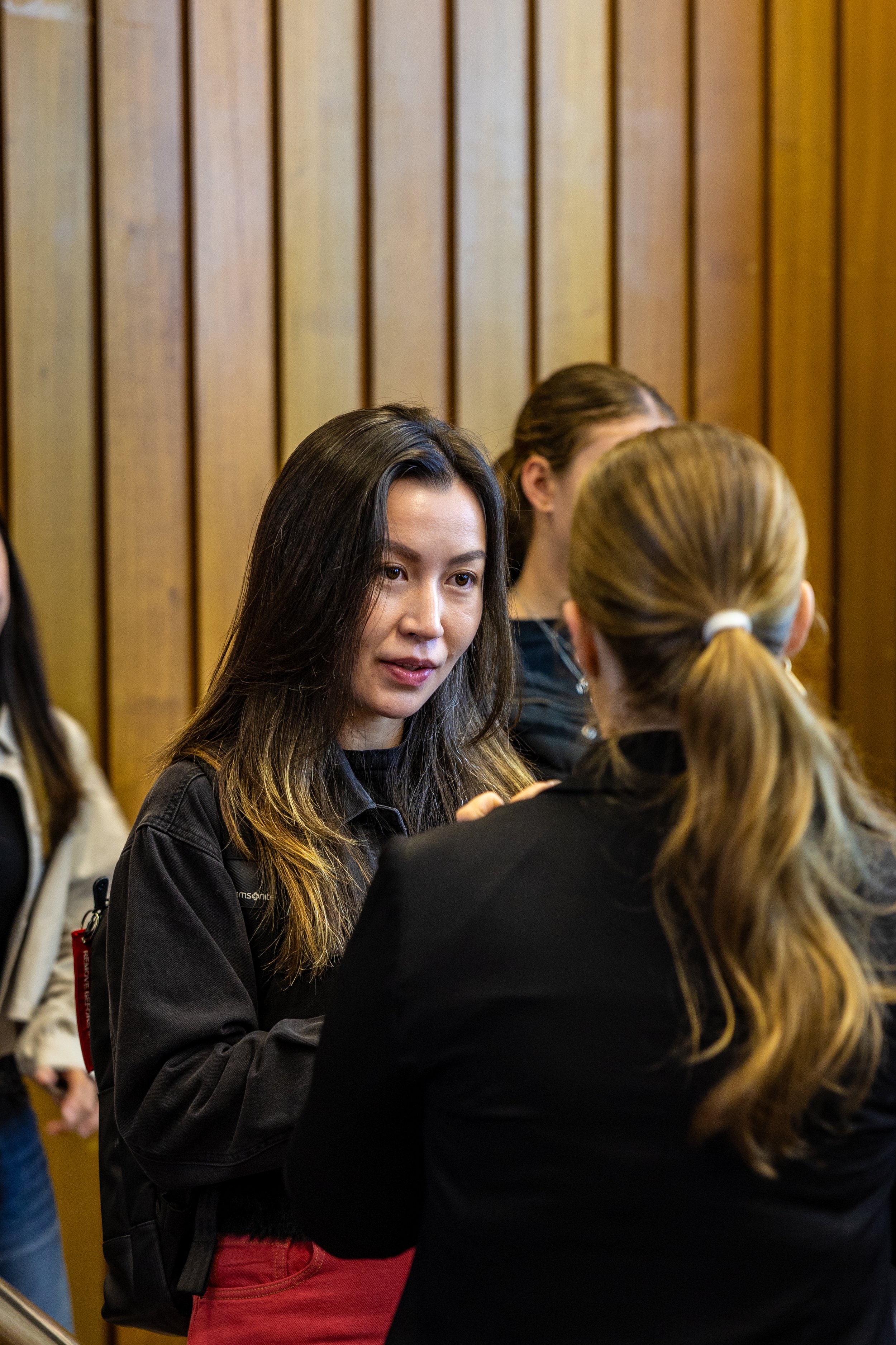 Two women engaged in conversation in a room with wooden paneling, one woman with black hair and casual clothing, the other with blond hair tied back.