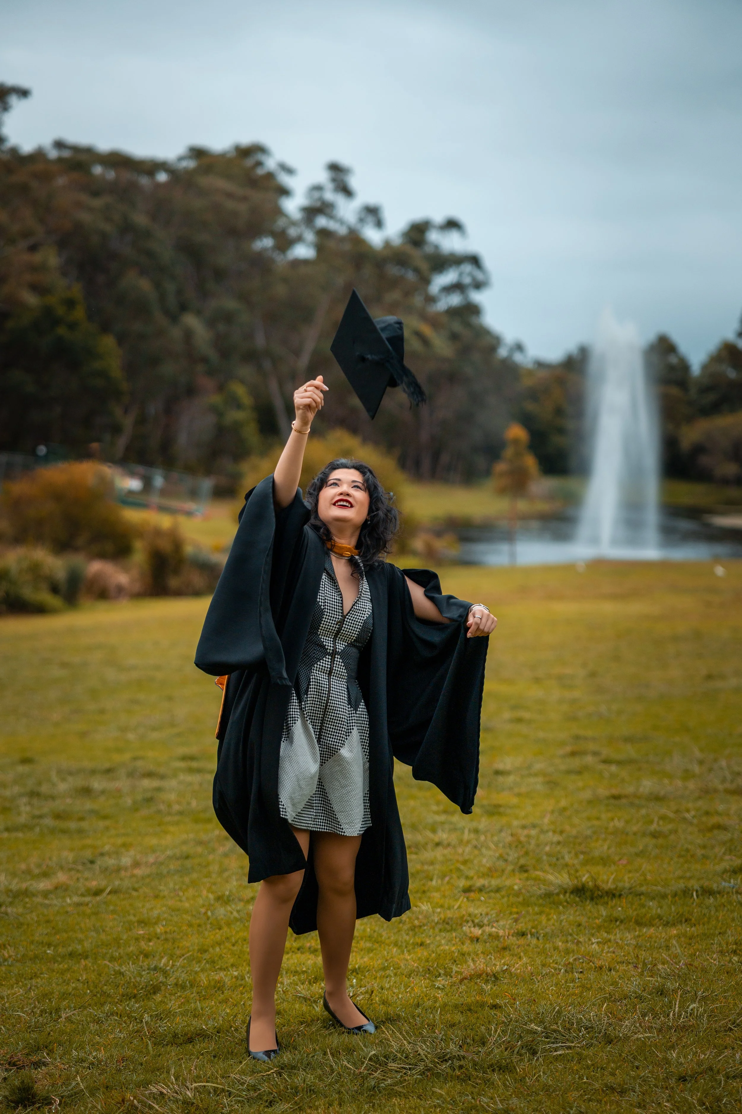 A woman in a graduation gown and cap throwing her cap in the air outdoors near a pond and fountain.