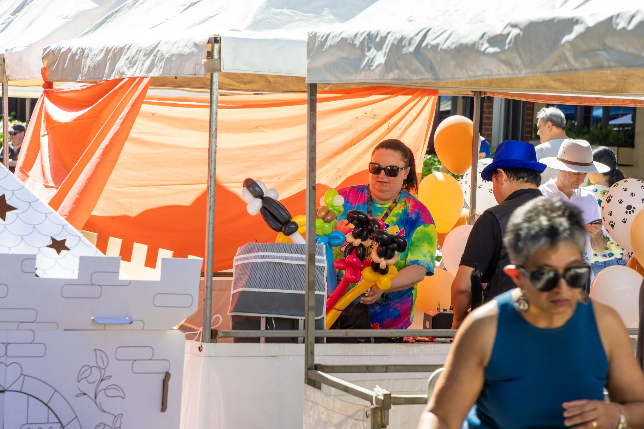 A woman in sunglasses and a colorful tie-dye shirt inside a vendor booth with orange tarps, surrounded by balloons and other people at an outdoor event.