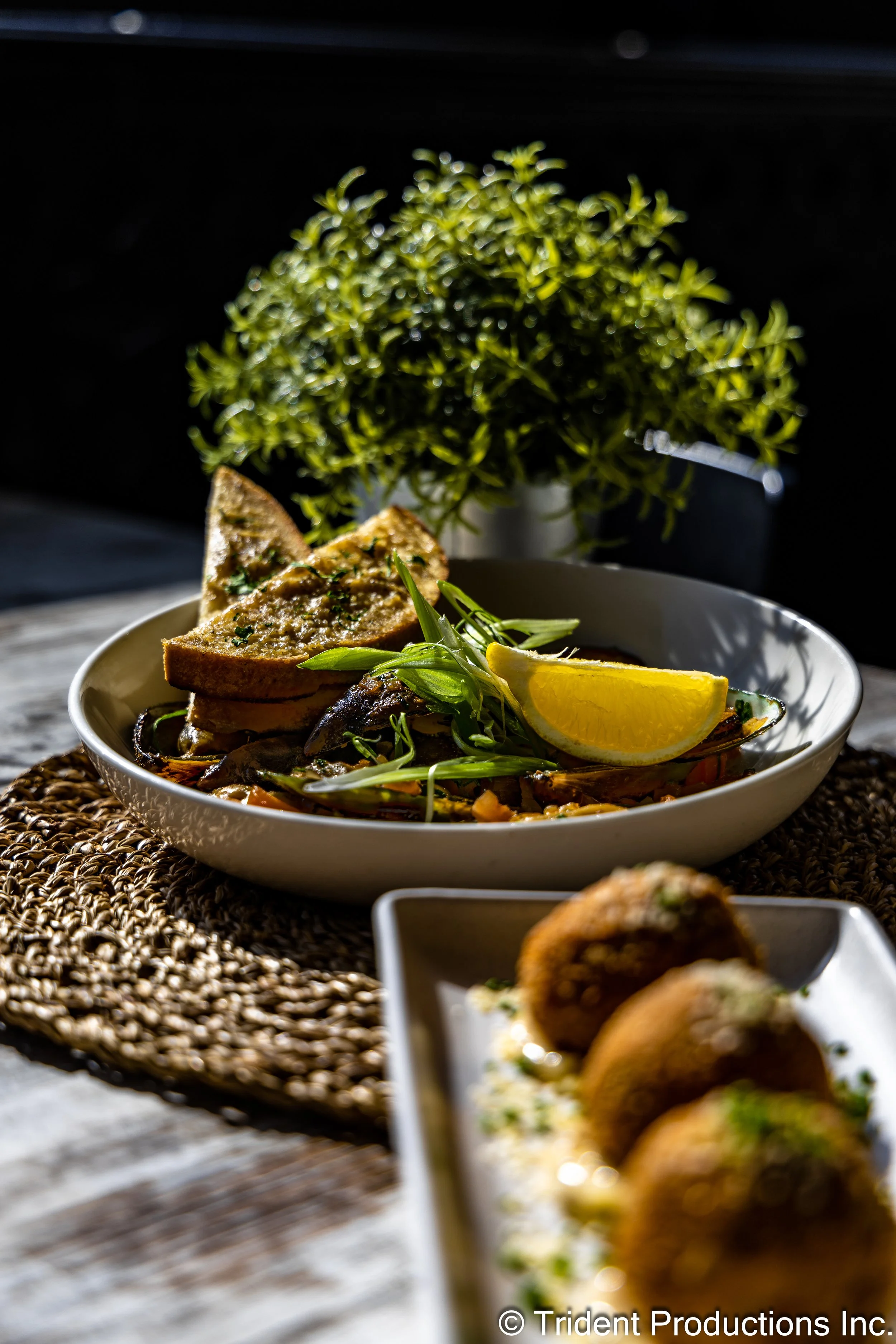 A plated meal with garlic bread, a lemon wedge, and seafood, garnished with herbs, with more seafood appetizers in the foreground.