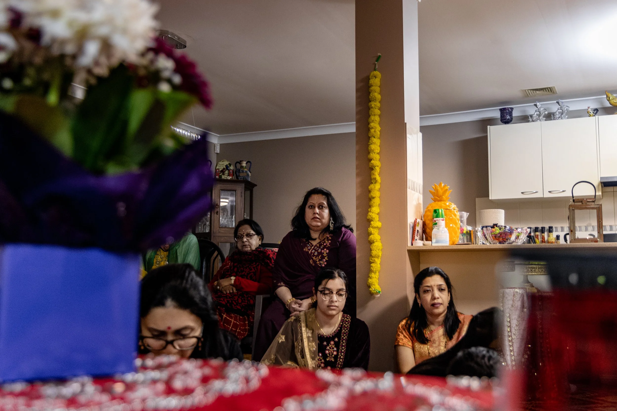 Women sitting and standing in a home with a kitchen in the background, decorated with yellow flowers and a pineapple-shaped decoration.