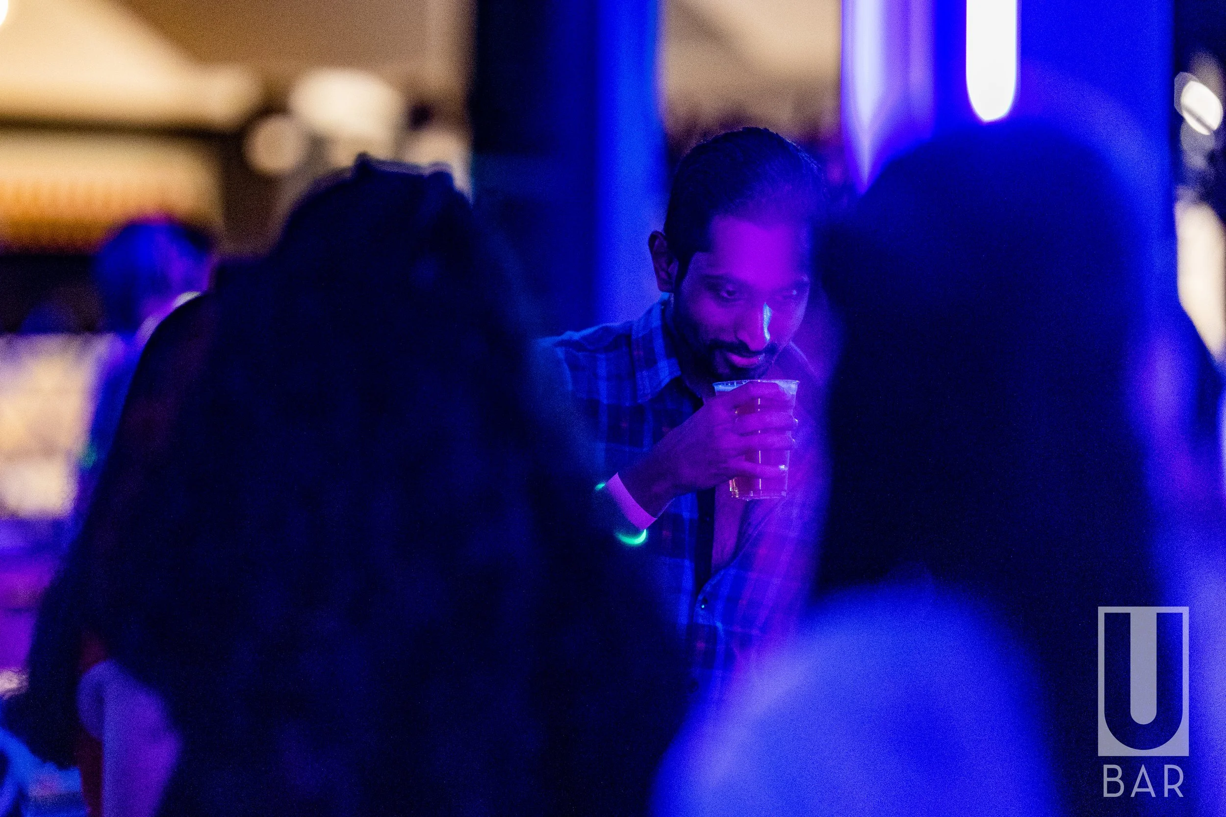 Man in plaid shirt holding drink in a dark, blue-lit bar with people around him.