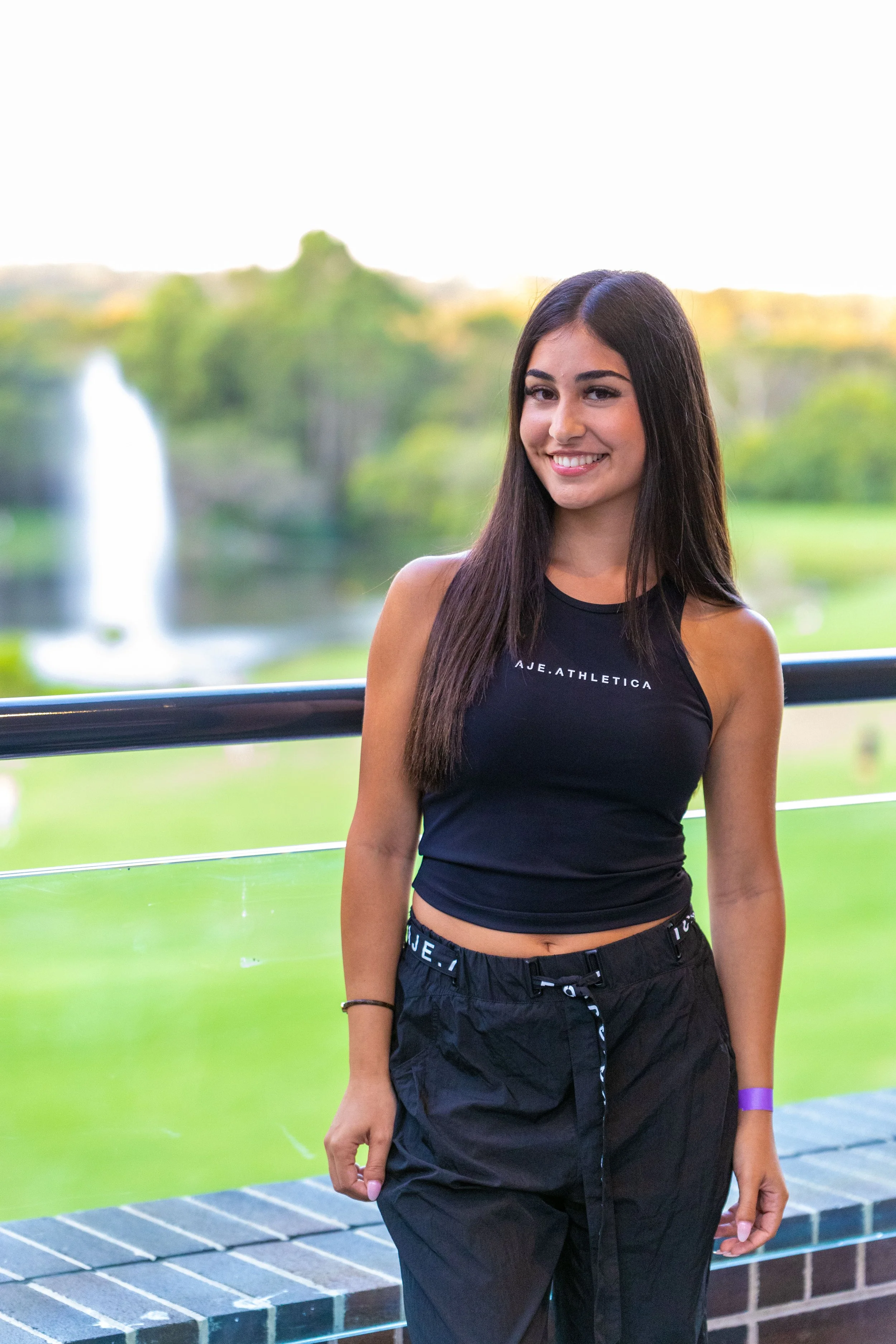 A young woman with long dark hair smiling outside near a lake with a fountain in the background.