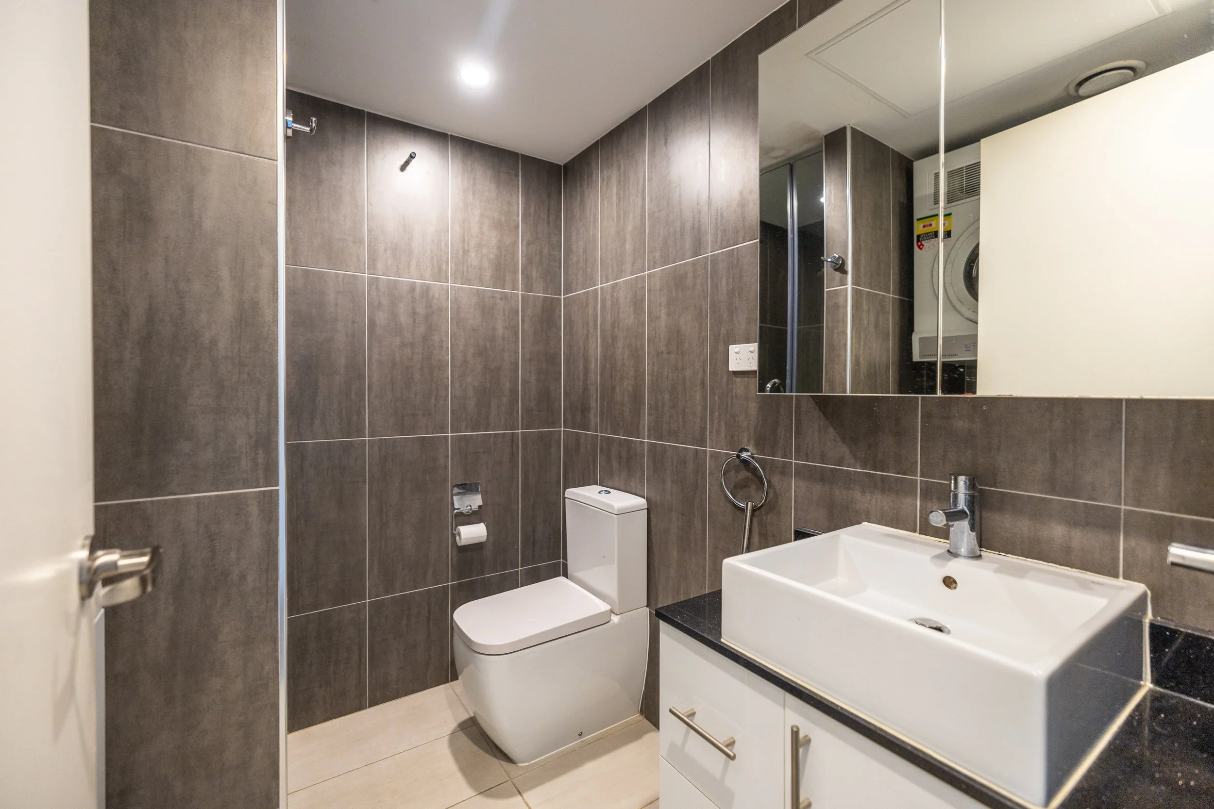 Modern bathroom with grey tiled walls, a white toilet, a white sink with black countertop, mirror, towel ring, and stacked washer and dryer