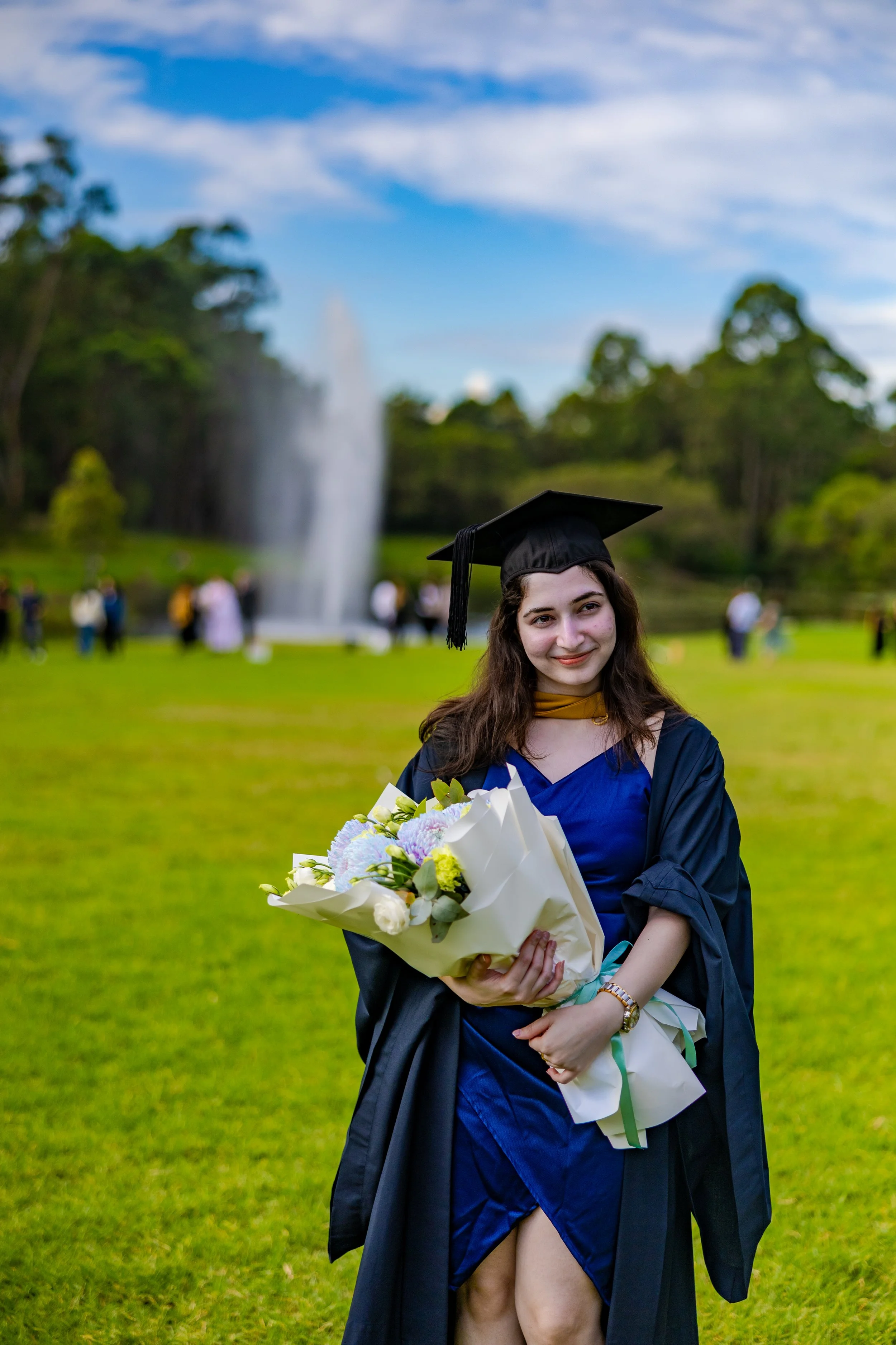 A young woman in graduation attire holding a bouquet of flowers at an outdoor graduation ceremony with a fountain and trees in the background.