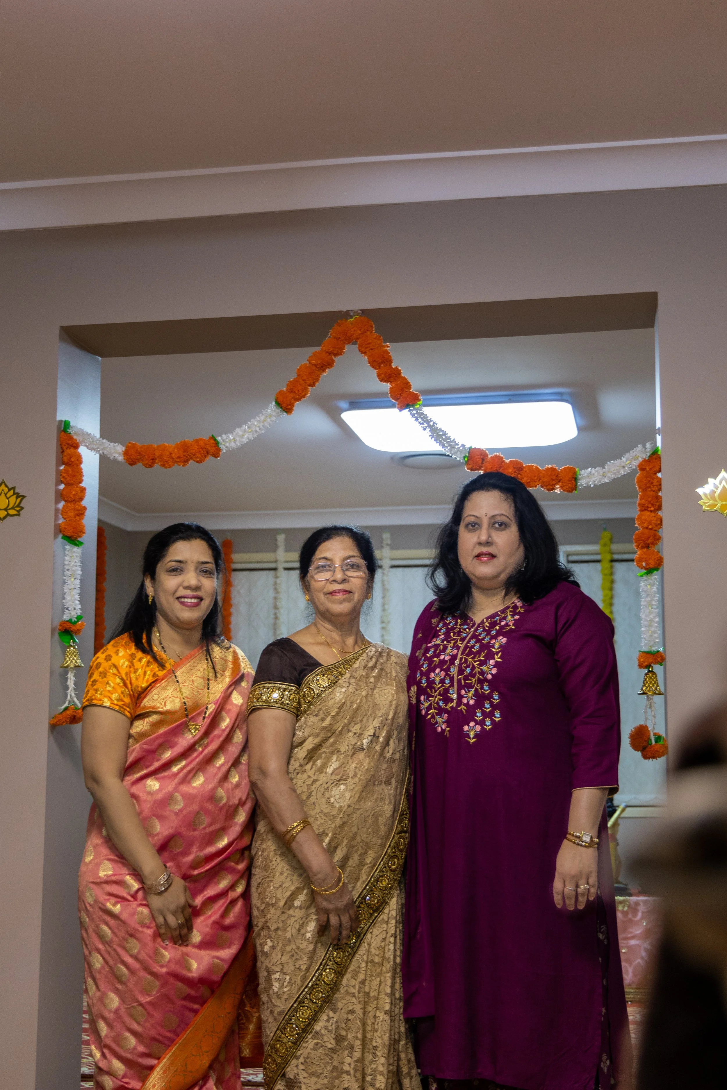 Three women dressed in colorful traditional Indian attire, standing together at a festive celebration with orange and white flower garlands and decorations in the background.