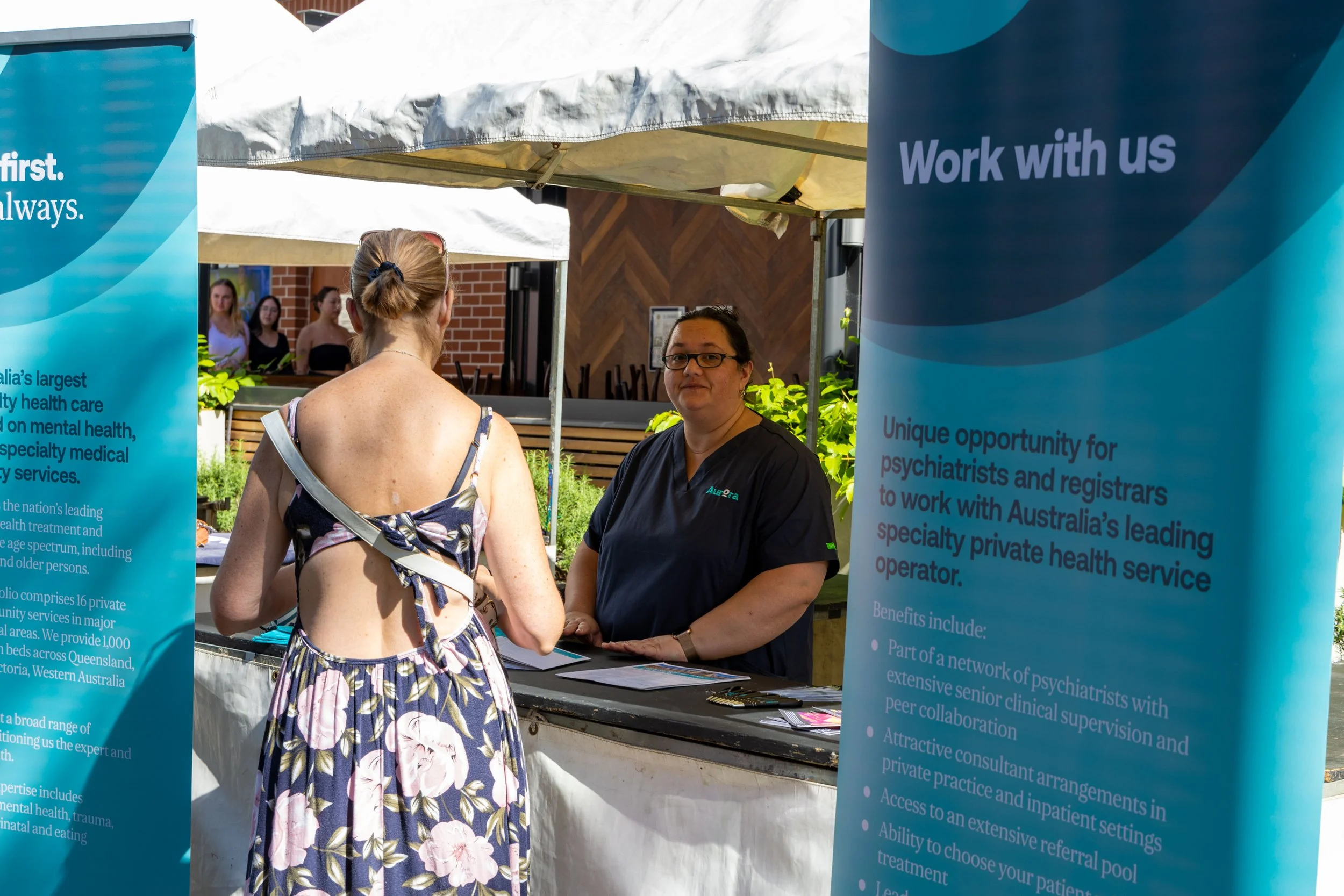 A woman in a floral dress with an open back talking to a healthcare worker at an outdoor booth with blue banners promoting a mental health service.