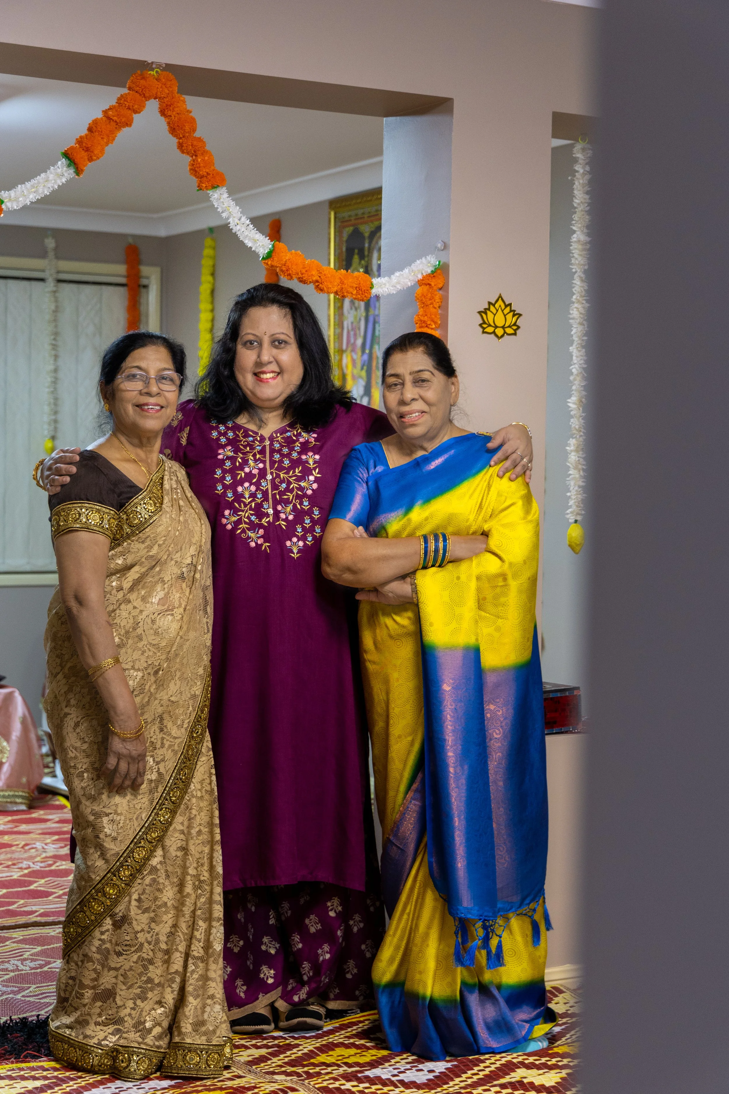 Three women in traditional Indian sarees and kurta standing together, smiling at a celebration decorated with marigold and white flower garlands.