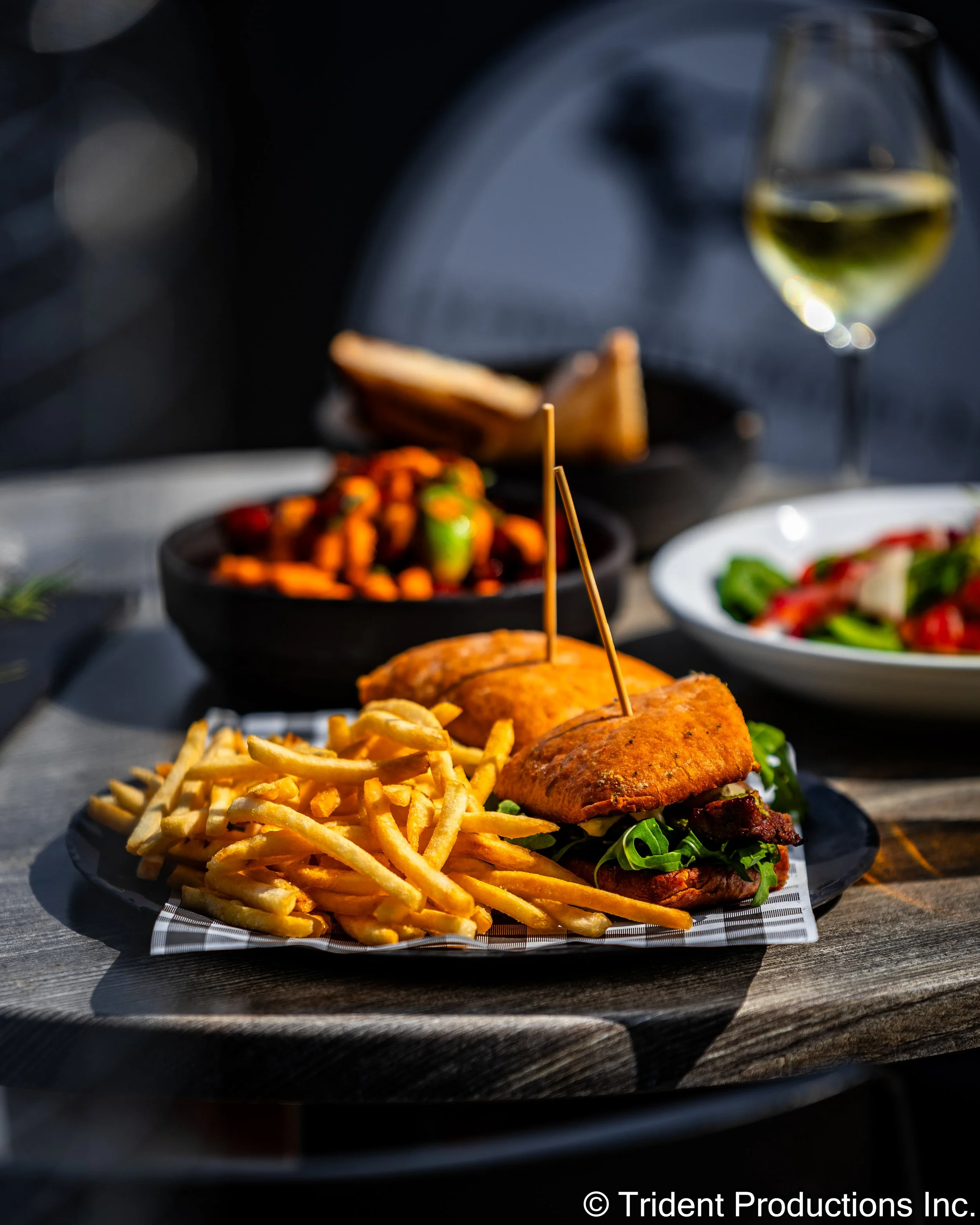 A plate with a fried fish sandwich with fries, a bowl of mixed vegetables, and a glass of white wine on a wooden table.