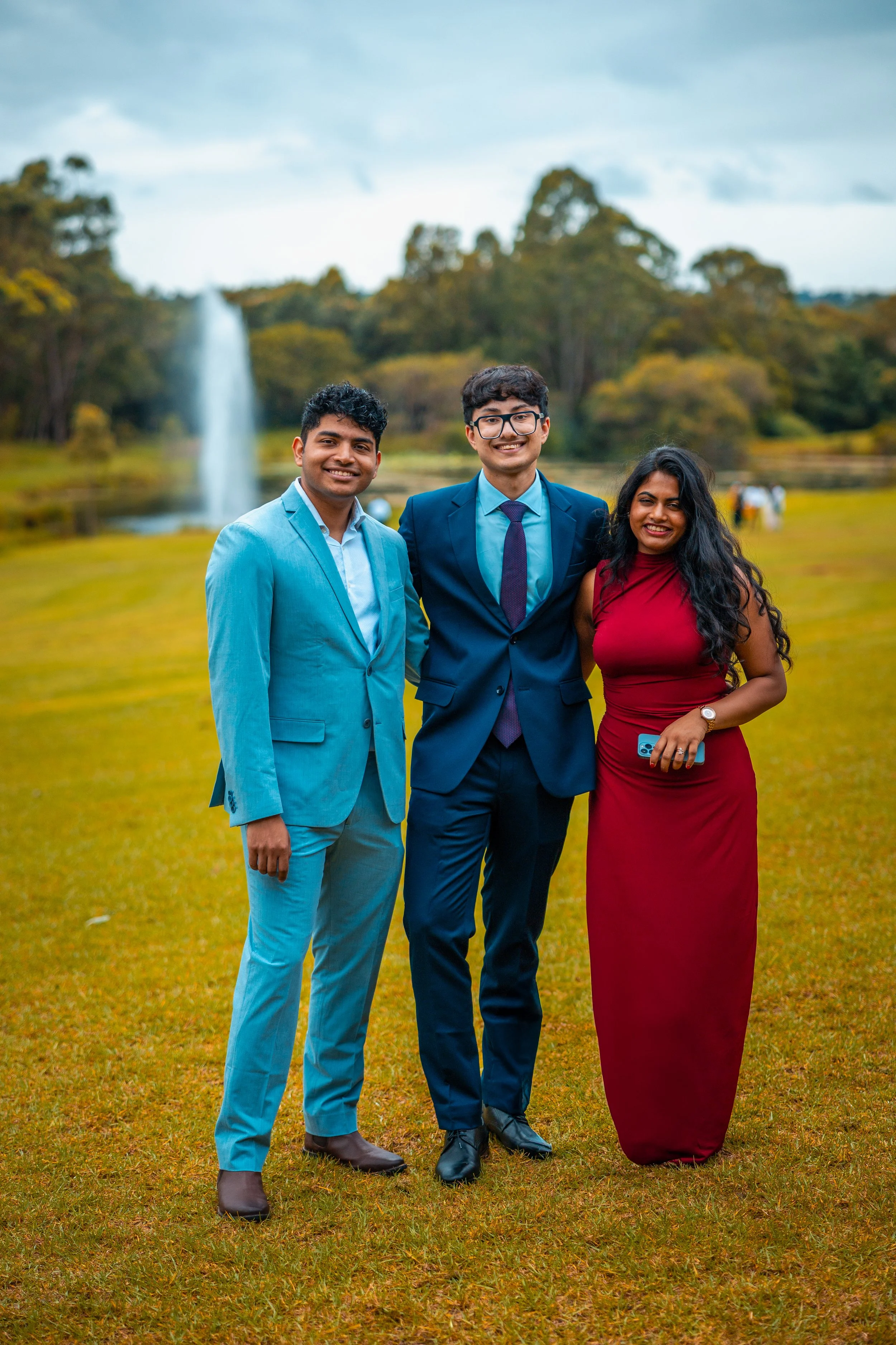 Three young adults in formal attire standing on grass in an outdoor park with trees and a fountain in the background, smiling at the camera.