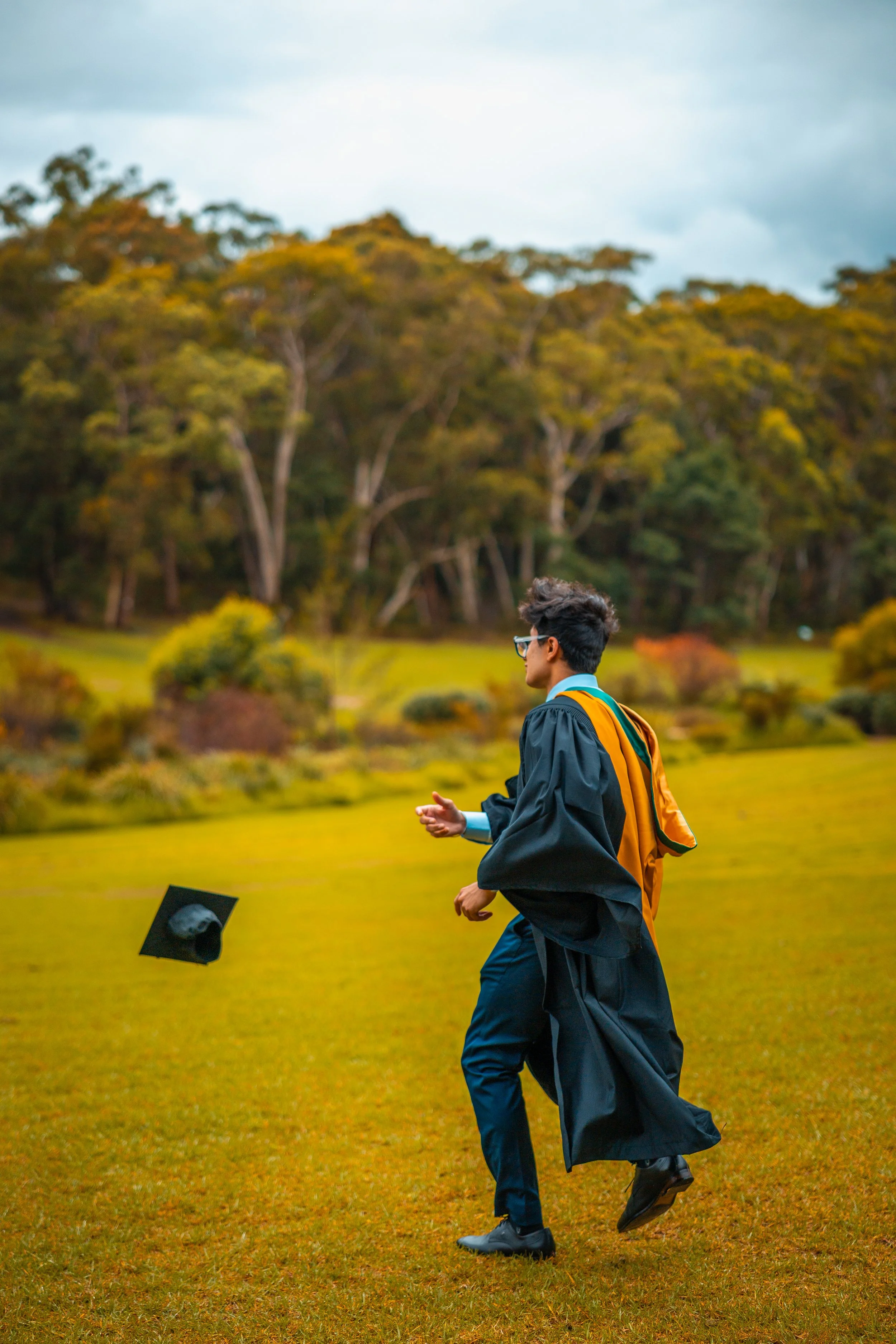 A young man in graduation attire, tossing his cap in the air on a grassy field with trees and a cloudy sky in the background.