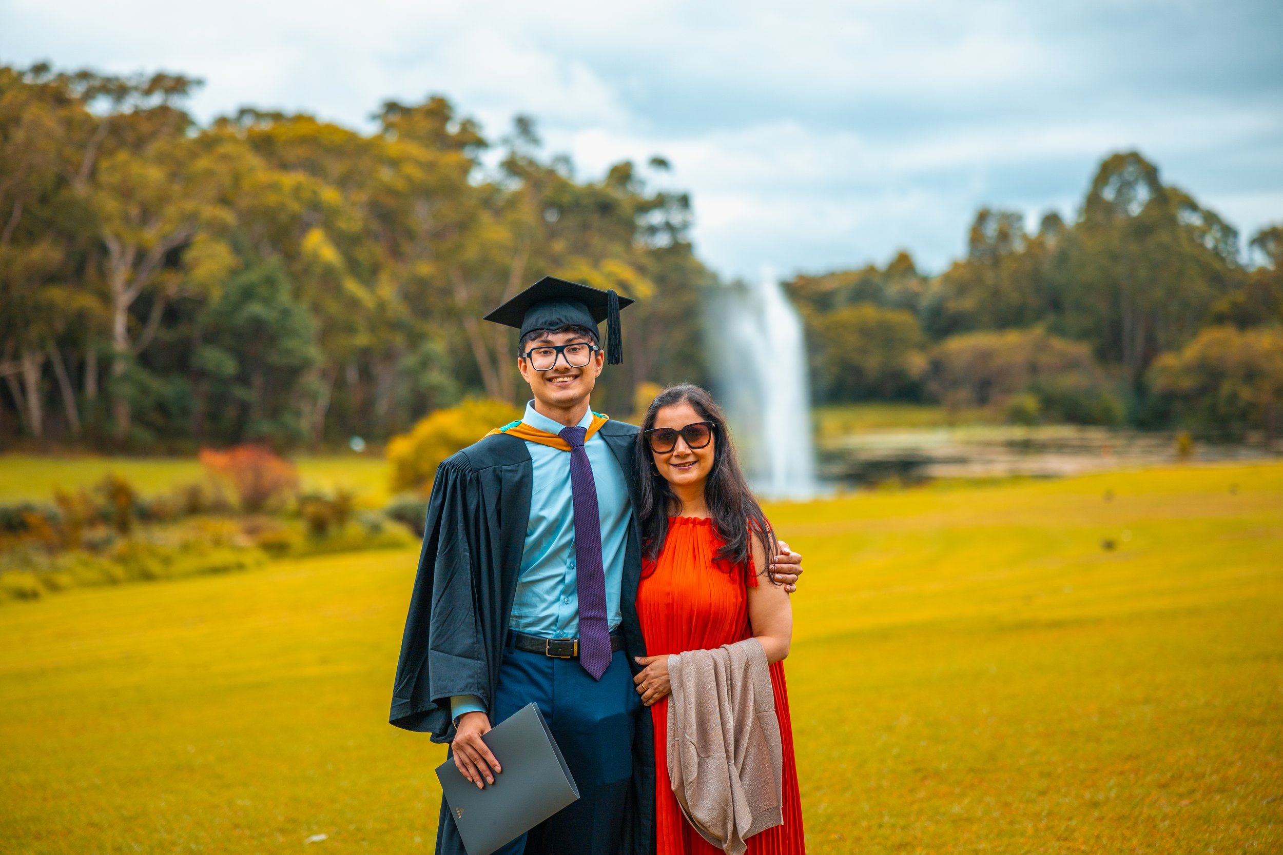 A young man in graduation attire, wearing a cap and gown, standing next to a woman in a red dress outdoors with a fountain and trees in the background.