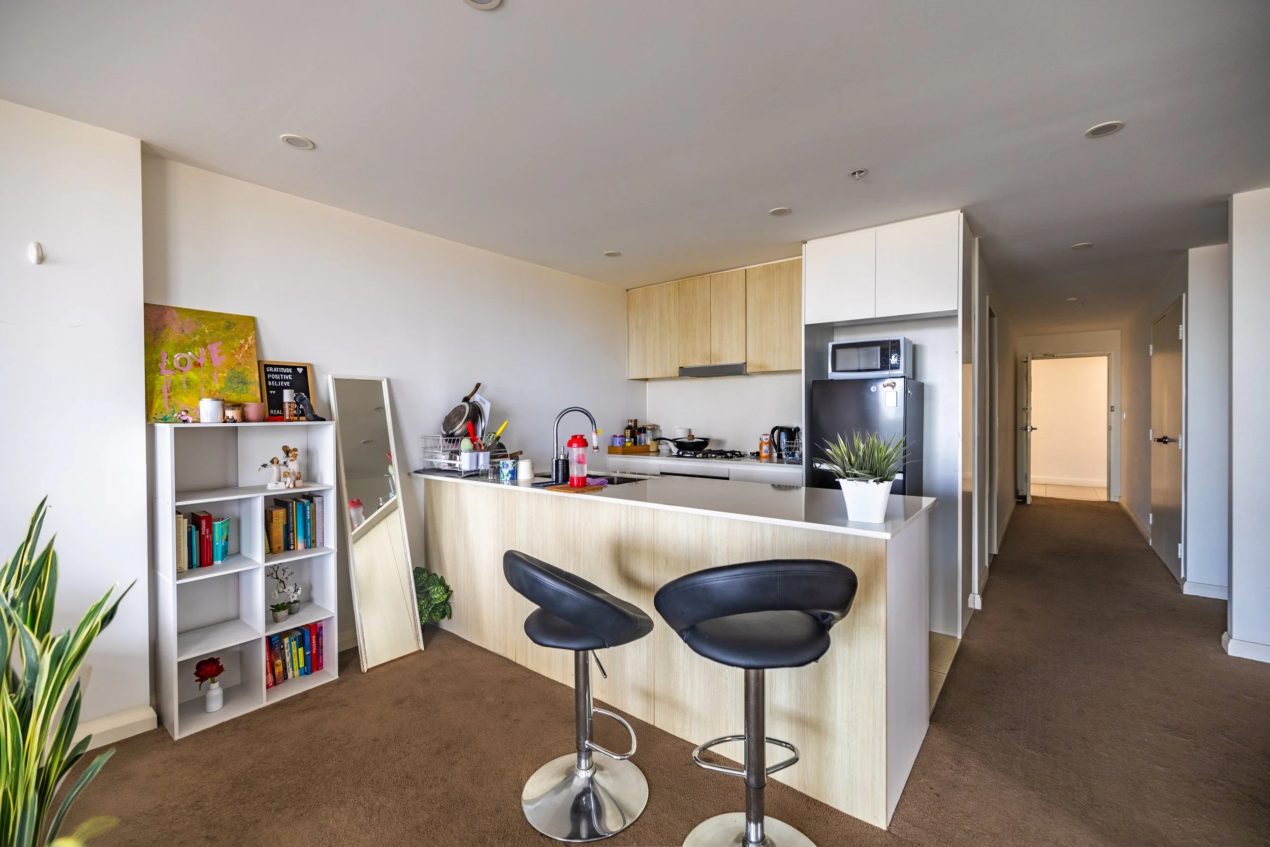 A modern kitchen with a breakfast bar, black stools, a fridge, microwave, and potted plant, with a hallway leading to other rooms.