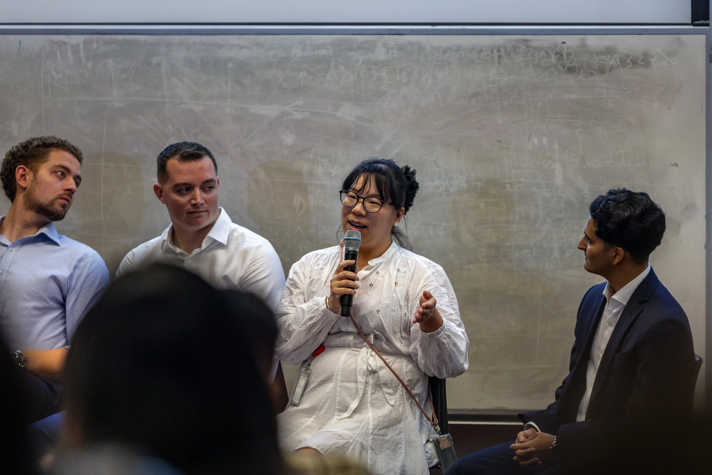 A woman speaking into a microphone during a panel discussion, seated between three men in business attire at an indoor event.